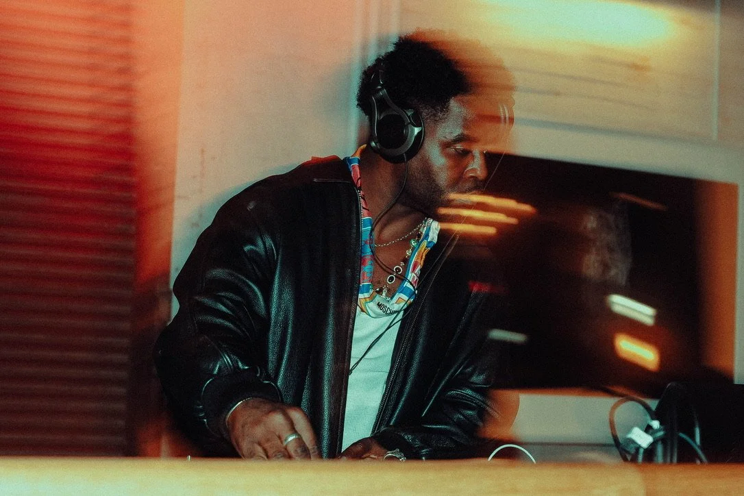A man with curly hair wearing headphones and a black leather jacket, working as a DJ in a dimly lit room.