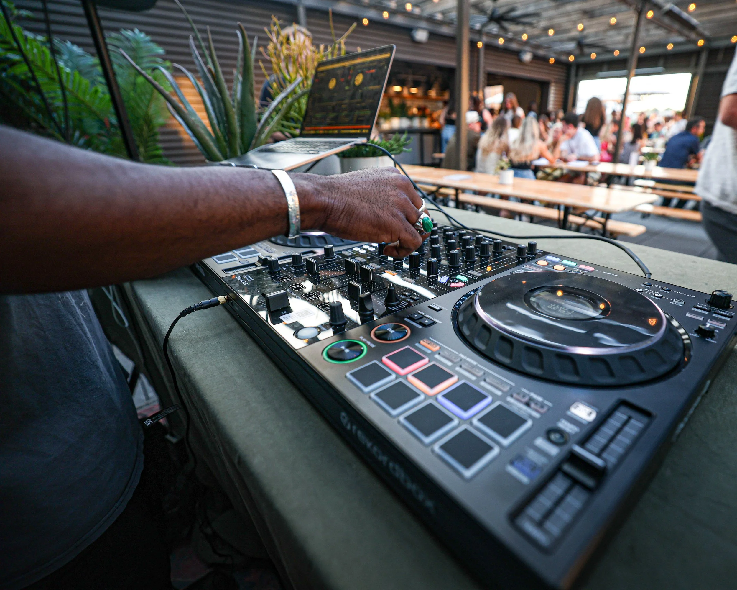 A DJ is mixing music at an outdoor event, with a turntable and laptop on the table surrounded by potted plants, while people are gathered and socializing in the background under string lights.