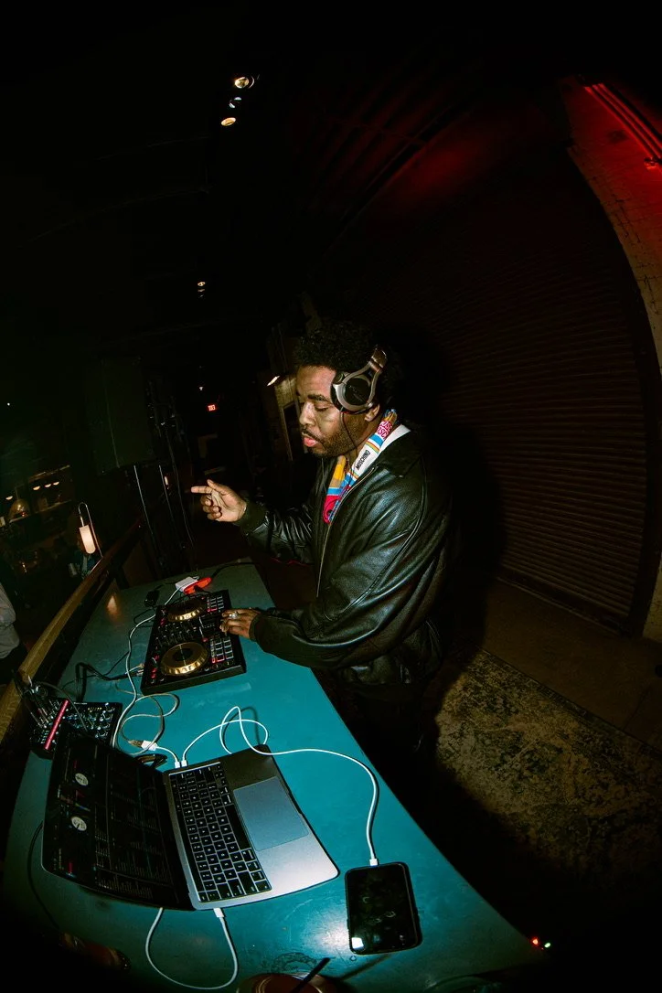 A male DJ with headphones mixing music on a DJ controller in a dimly lit club, with laptop and smartphone on the table.