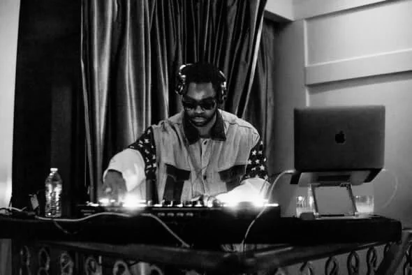 A person wearing sunglasses and headphones, standing behind a DJ mixing console with a laptop and water bottle, in a room with curtains.