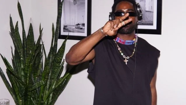 A man wearing sunglasses and jewelry making a peace sign with his fingers, standing next to a potted plant indoors.