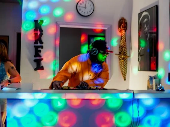 DJ wearing headphones, performing at a colorful illuminated bar with lights, a clock, and artwork on the walls.