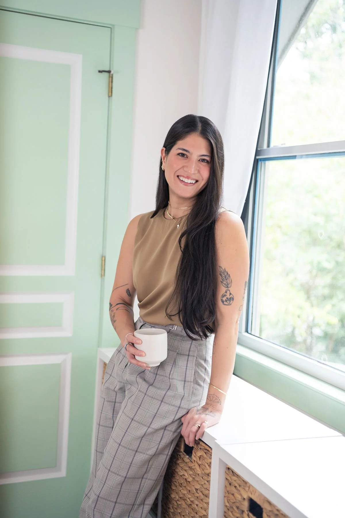 A woman with long dark hair, wearing a beige sleeveless top and plaid pants, smiling while holding a mug, standing near a window inside a light-colored room with green and white-tiled walls.