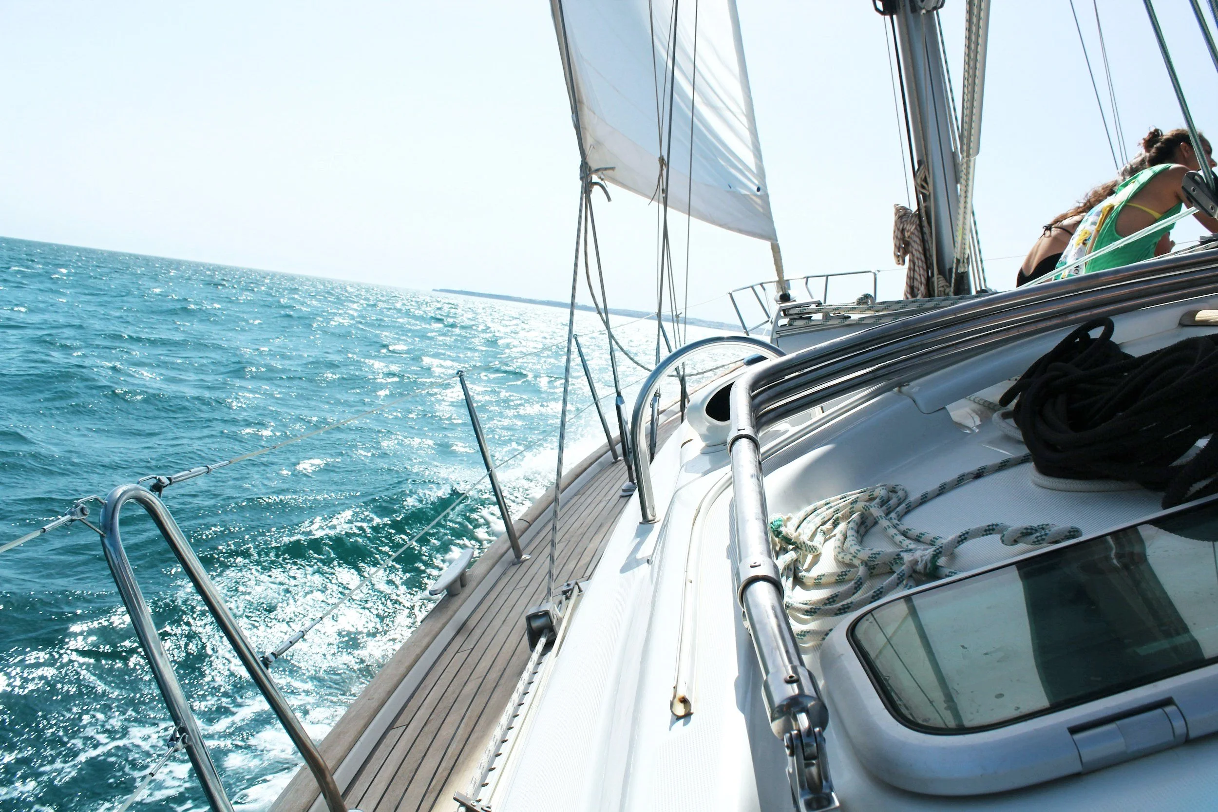 Sailboat navigating on an open sea with clear blue water and white sails, deck view showing ropes and railing, sunny day.
