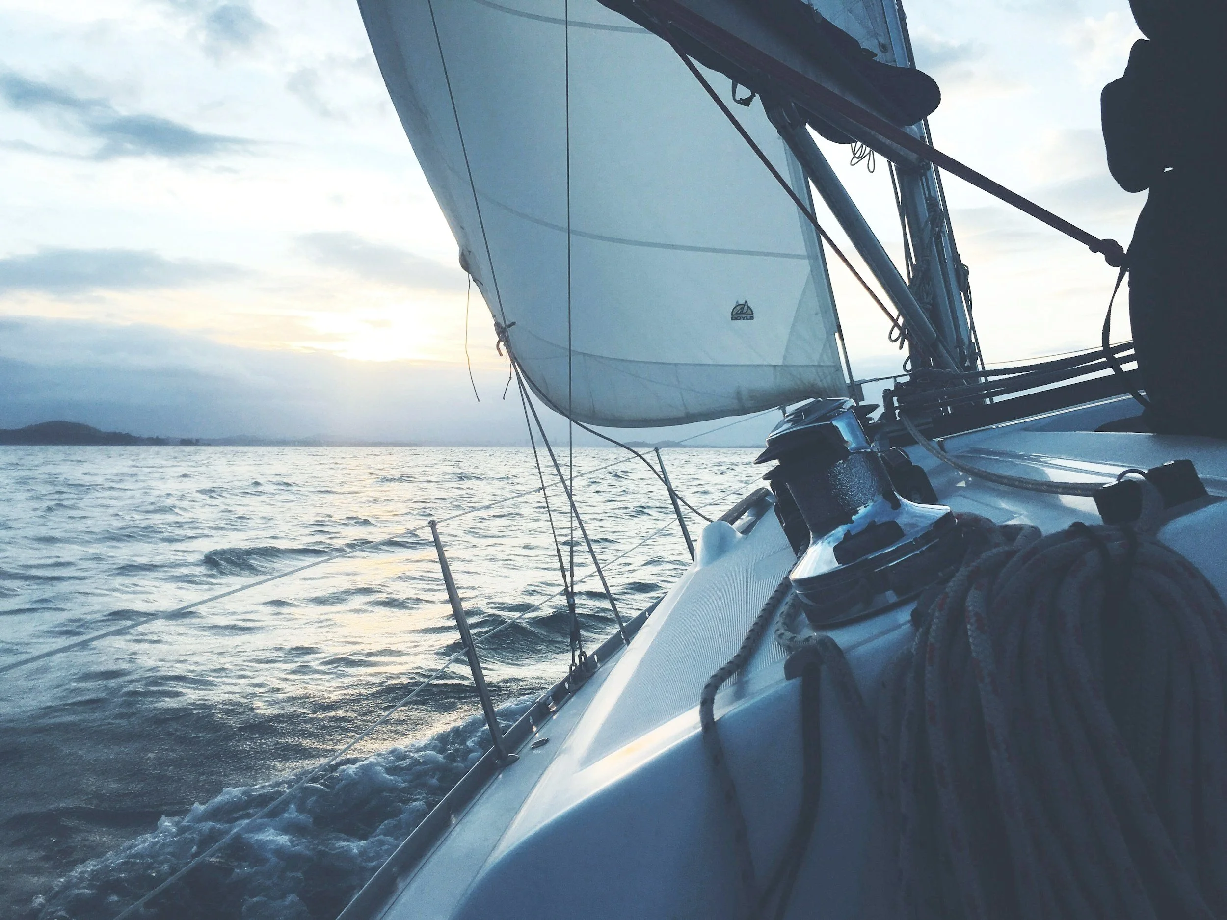 Close-up of a sailboat deck with ropes, winches, and sails in action on the water, at sunset.