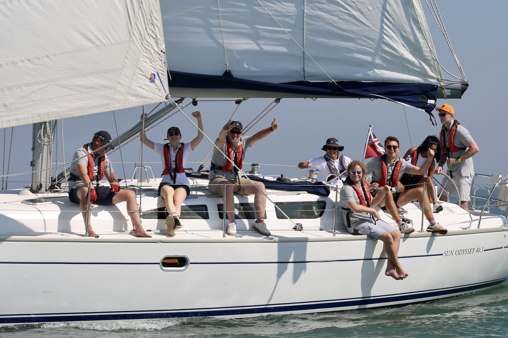 Group of people sailing on a boat, smiling and enjoying the sunny weather. The boat is a Sun Odyssey 40.3 model, and the passengers are wearing life jackets.