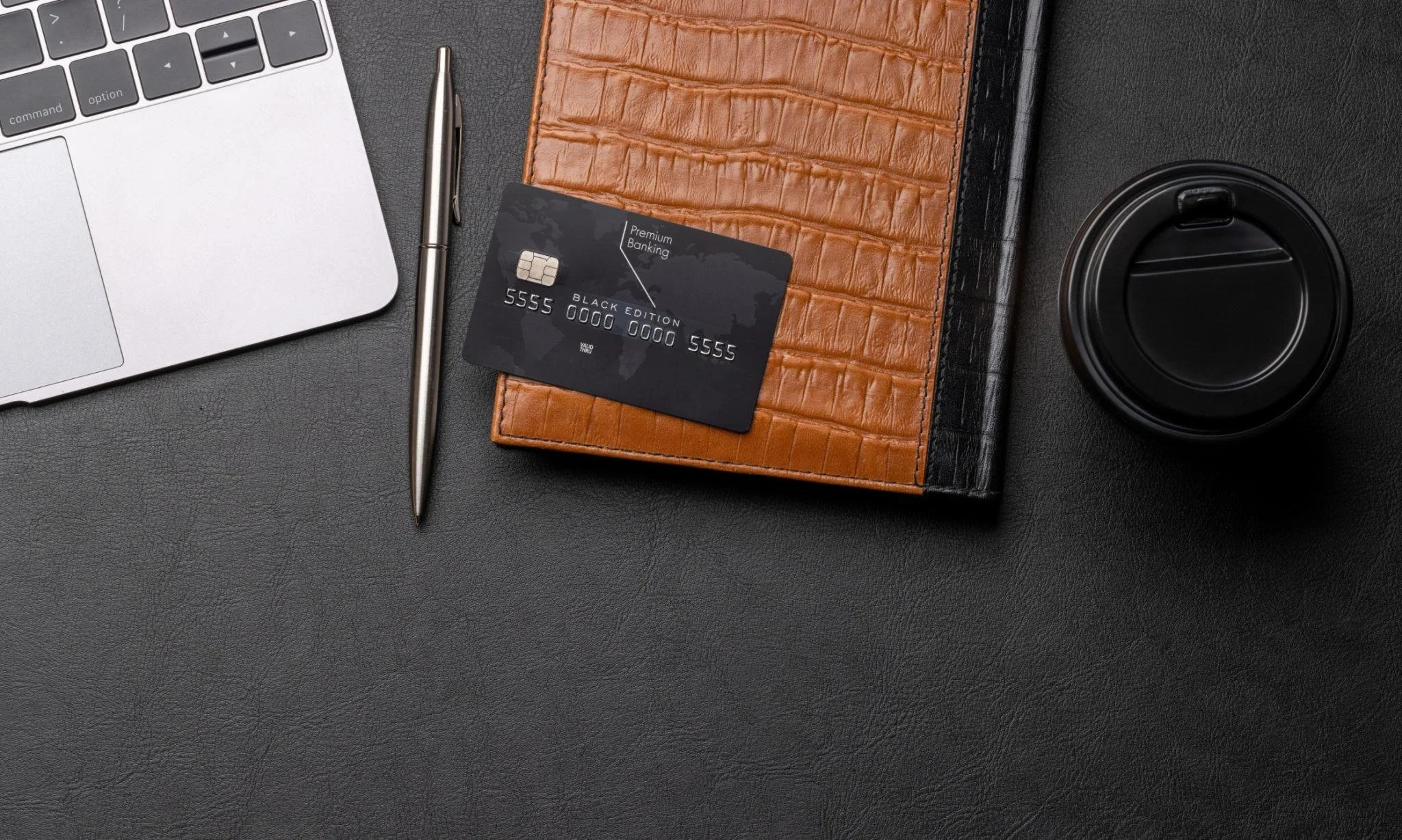 Close-up of a workspace with a silver laptop, a silver pen, a black premium banking card, a brown textured notebook or wallet, a black disposable coffee cup, all arranged on a black textured surface.