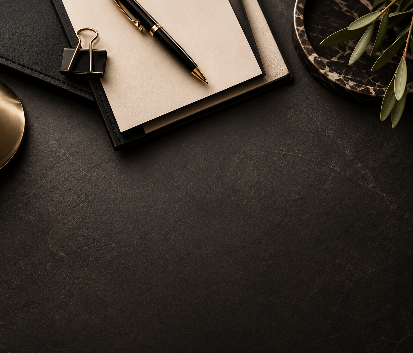 A black notebook with a gold clip, a black and gold pen on a white notepad, a marble dish with green leaves, and a corner of a gold object on a black textured surface.