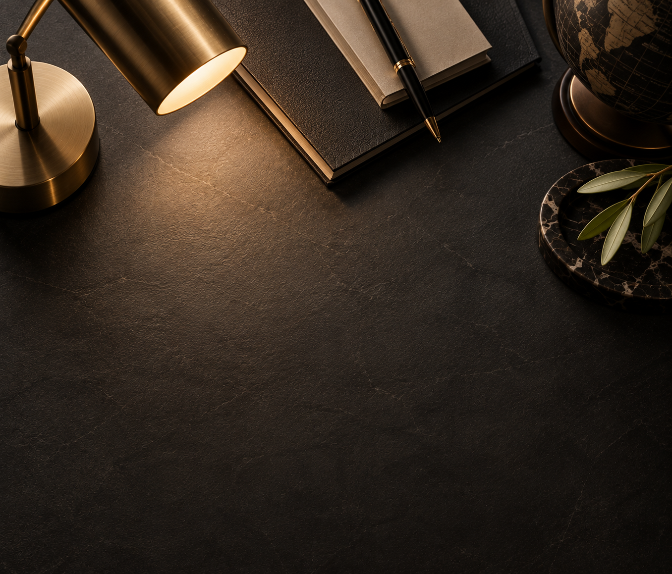 A dark textured desk surface with a gold desk lamp, a closed notebook, a pen, a globe, and a small plant on a marble tray.