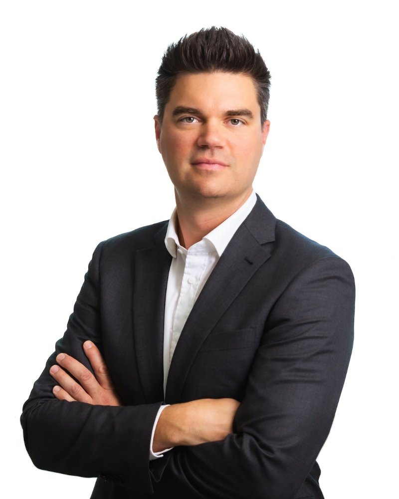 Seth Baker in a dark suit with a white shirt standing with arms crossed against a white background.