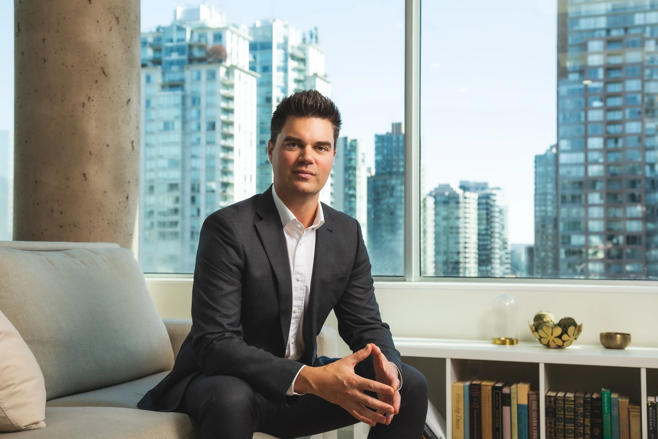 A man in a dark suit sitting on a beige sofa in a modern office with large windows showing a cityscape of tall buildings.