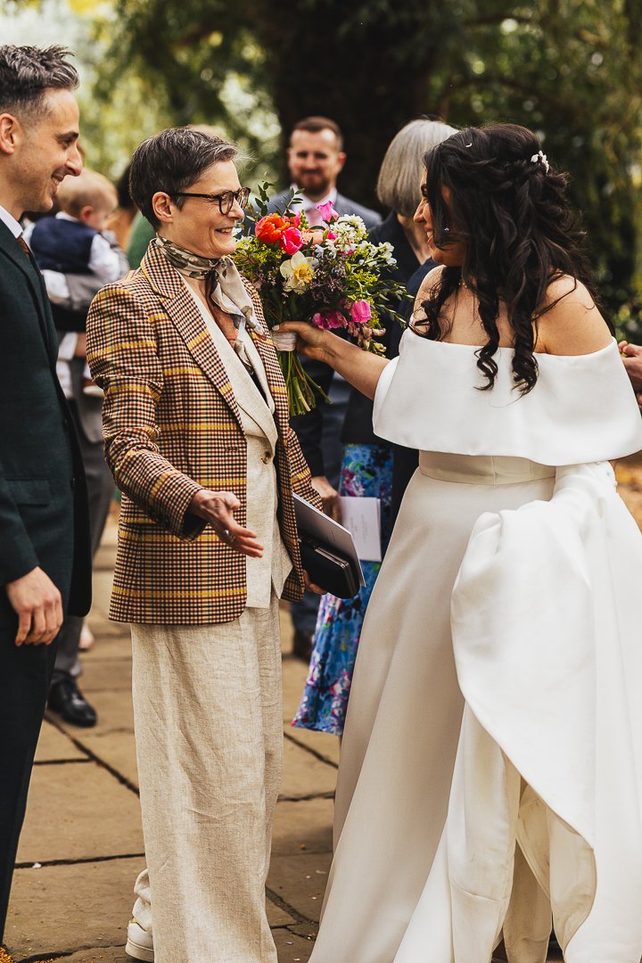 A group of people at a wedding, with one person in a white dress holding a colorful bouquet, outdoors with trees in the background.