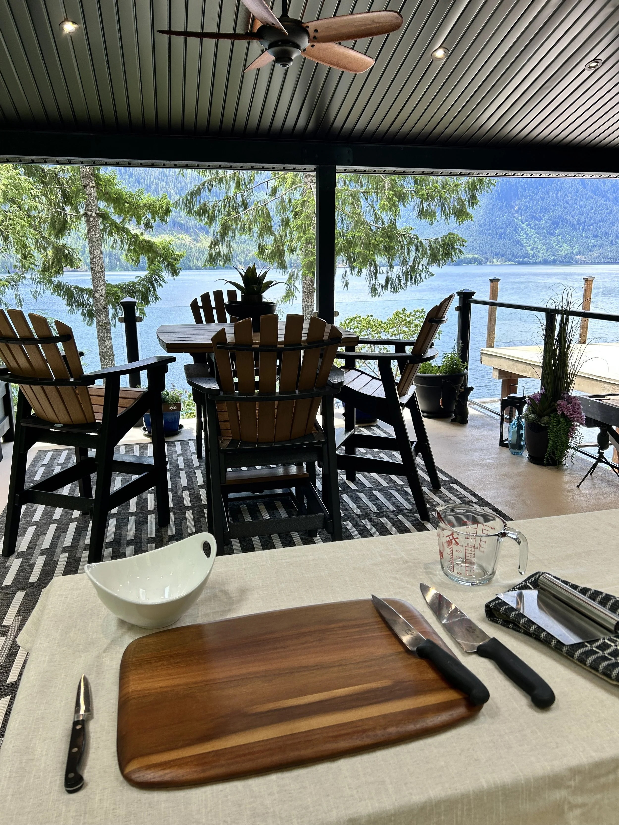 Dining table set with knives, a cutting board, a measuring cup, and a bowl, overlooking a lake with trees and mountains in the background, under a ceiling fan.