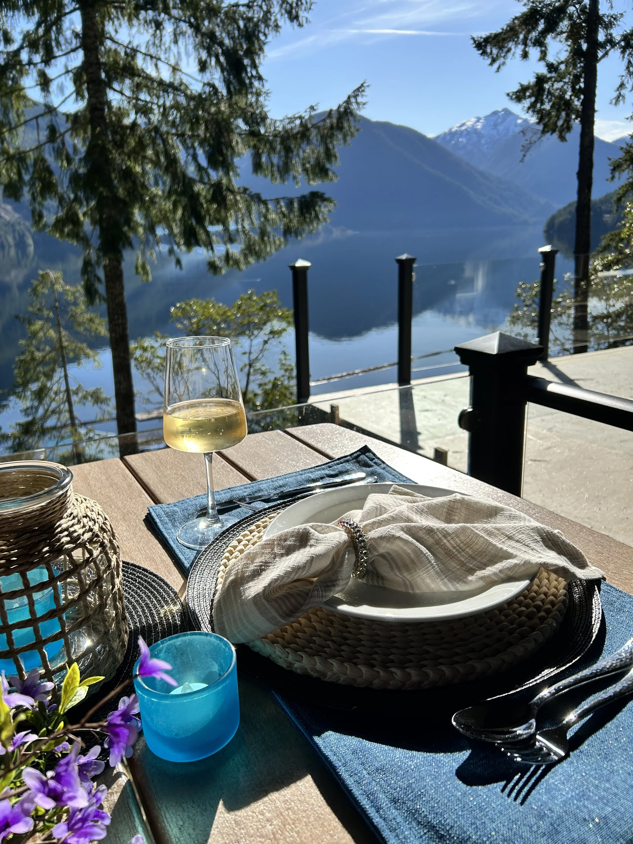 A lakeside outdoor dining scene with a glass of white wine, a plate with a napkin and jewelry, a woven basket, and purple flowers on a table, with a view of a calm lake, trees, and snow-capped mountains in the background under a clear blue sky.