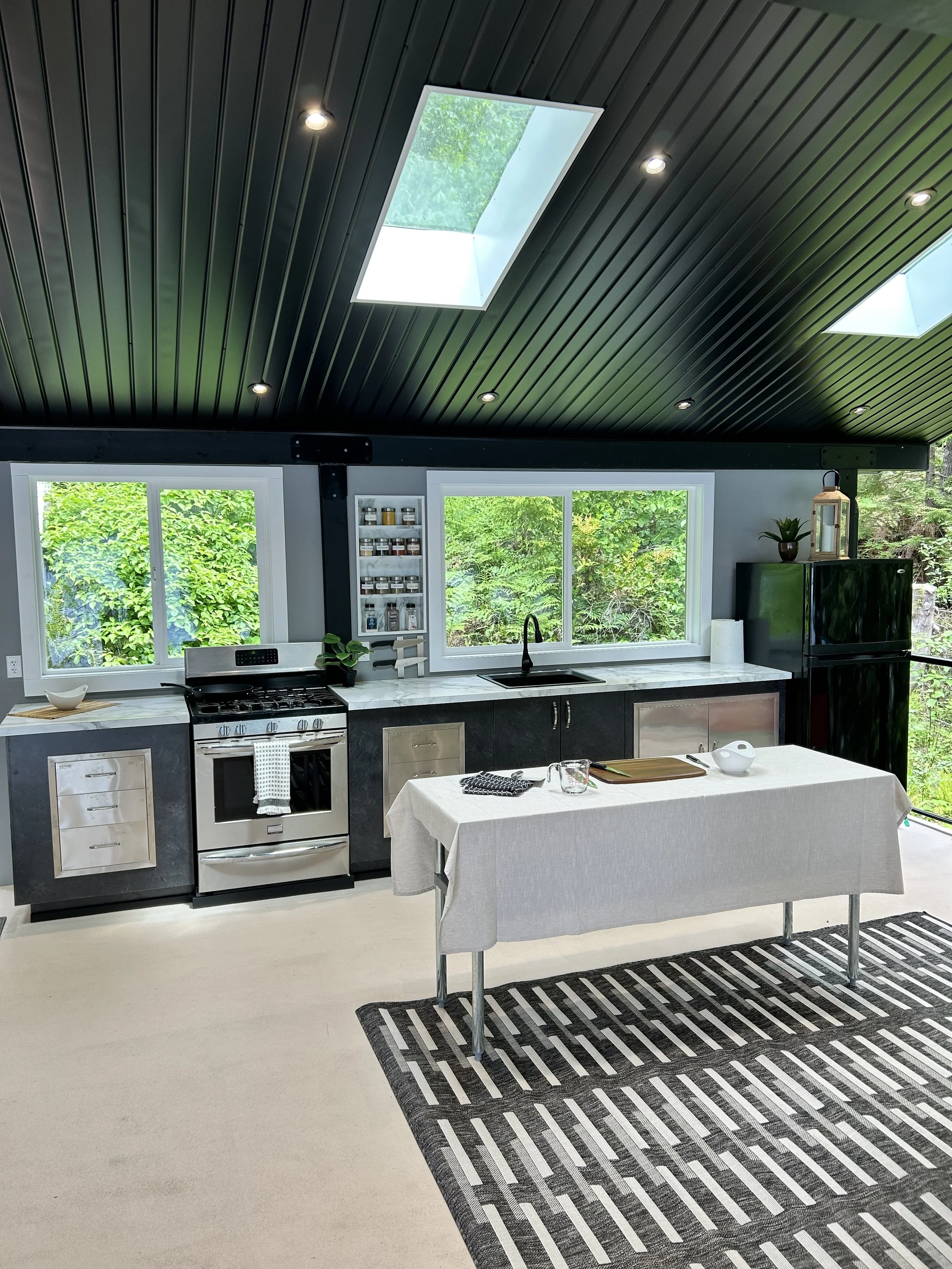 Modern kitchen with black cabinetry, stainless steel appliances, white marble countertops, large windows, and skylights in the black ceiling.