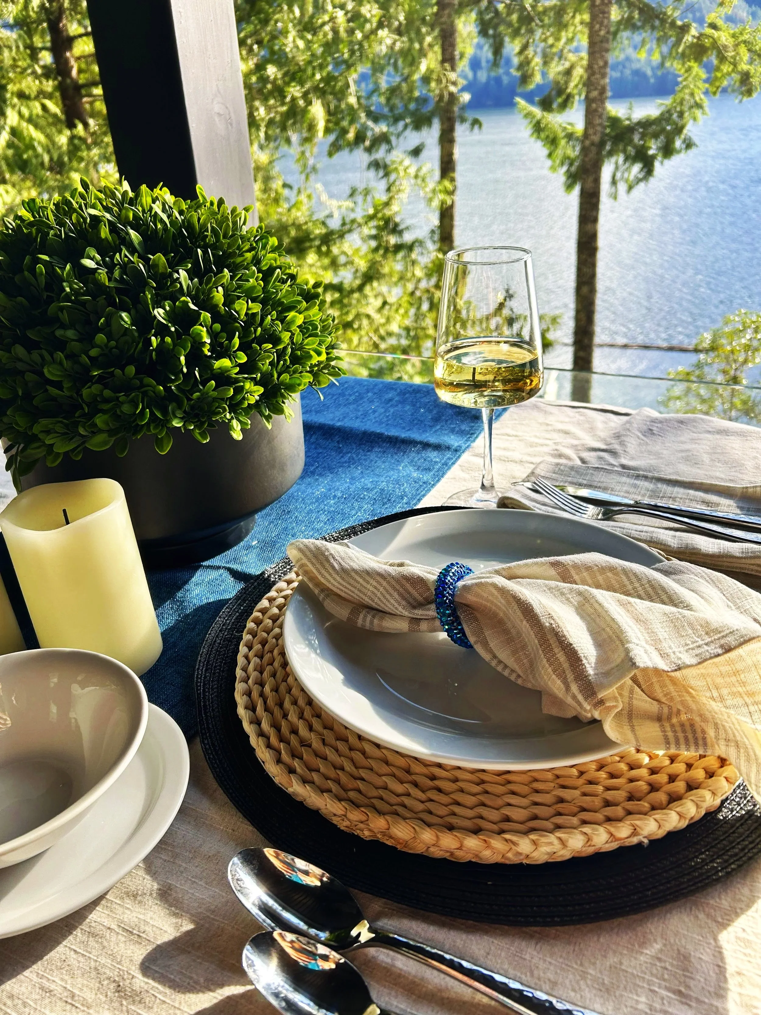 A table set for dining outdoors with a view of a lake and trees in the background. The table has a blue tablecloth, a potted green plant, a glass of white wine, a candle, a basket with a plate of food, and cutlery wrapped in a cloth napkin.