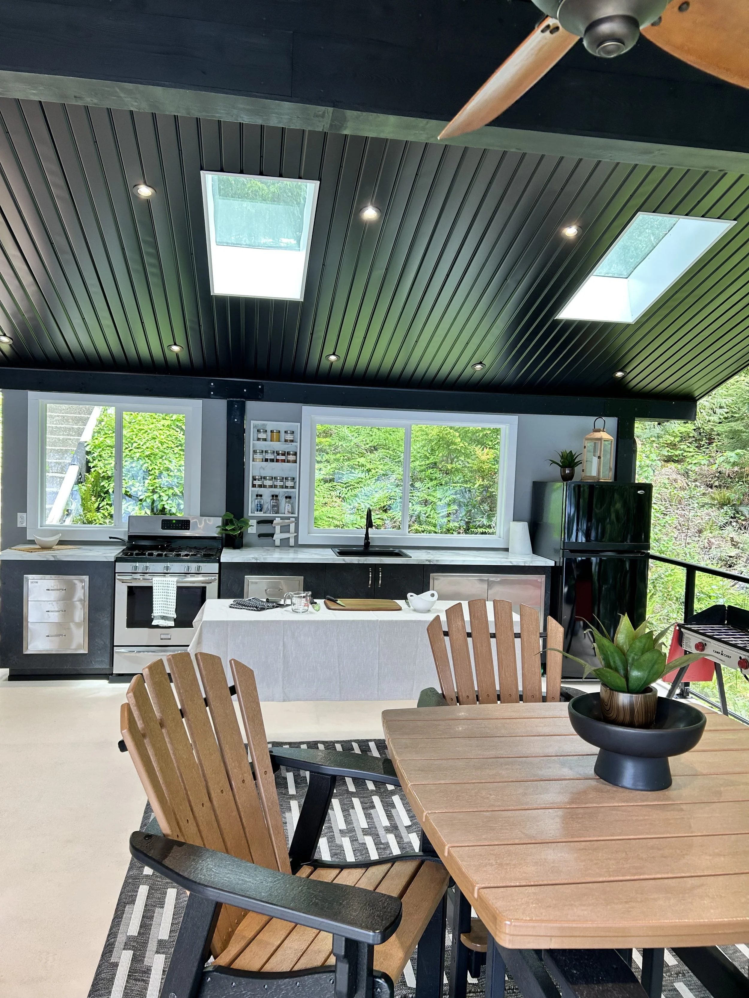 A modern kitchen with black ceiling panels, skylights, and large windows showing greenery outside. There is a wooden dining table with chairs and a potted plant, and black cabinetry with a white countertop, a black refrigerator, and stainless steel appliances.