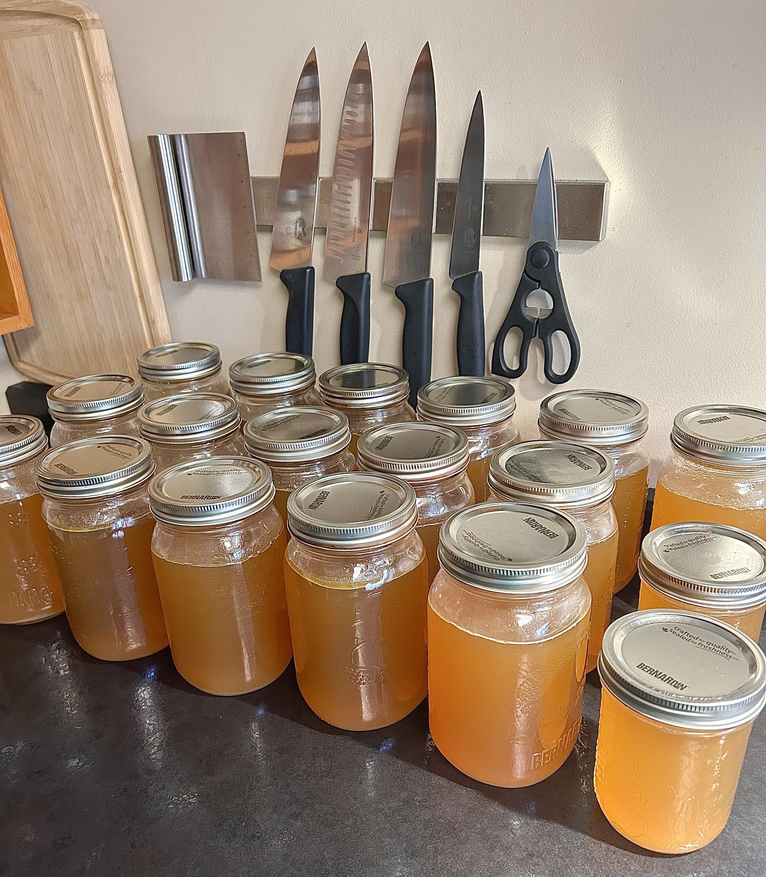 Multiple jars of house made chicken stock arranged on a black surface with a wall mounted knife rack holding six knives and a pair of scissors in the background.
