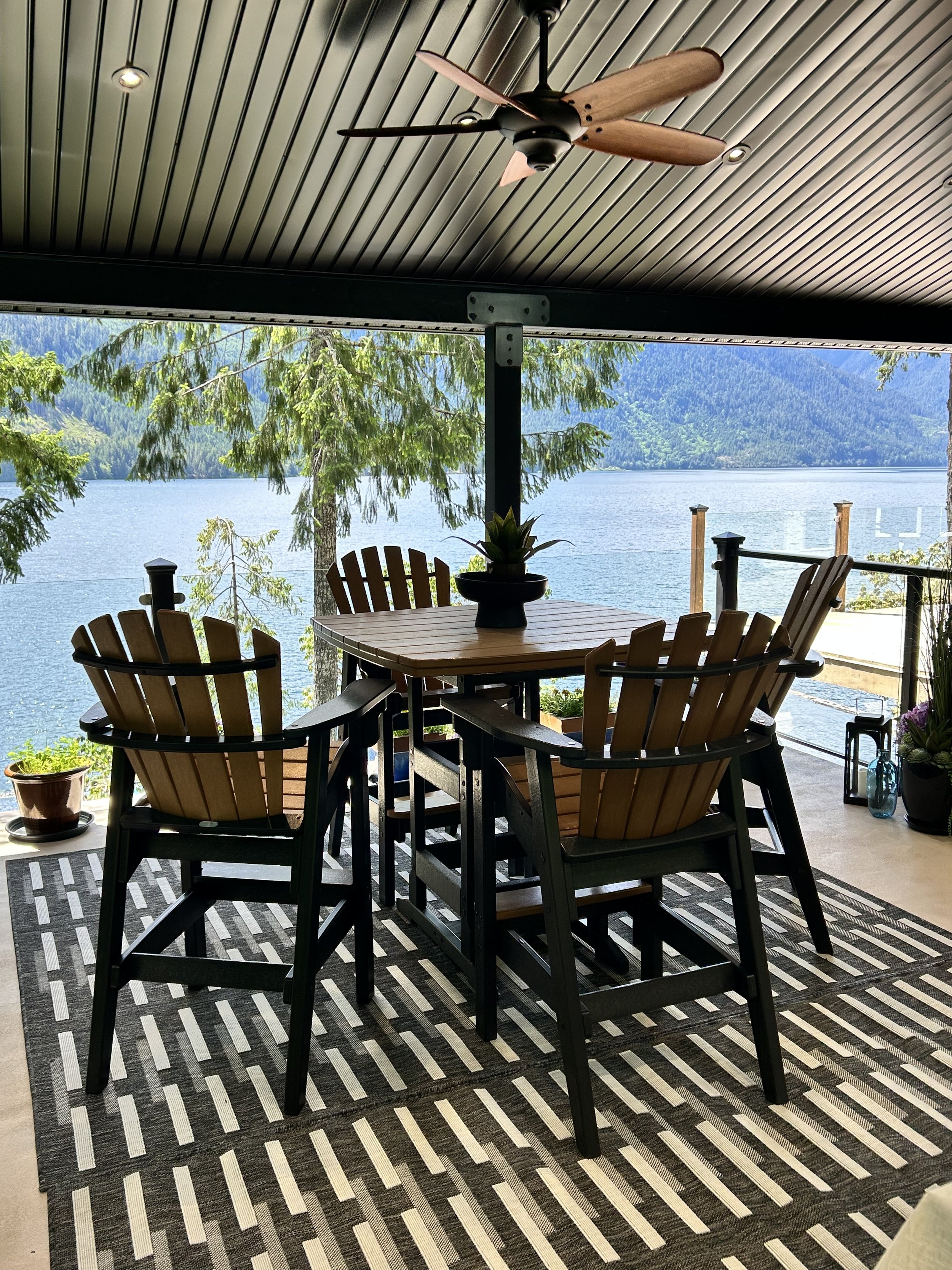 Indoor patio with a wooden dining table and six Adirondack chairs, overlooking a lake with pine trees and mountains in the background, ceiling fan overhead, and potted plants nearby.