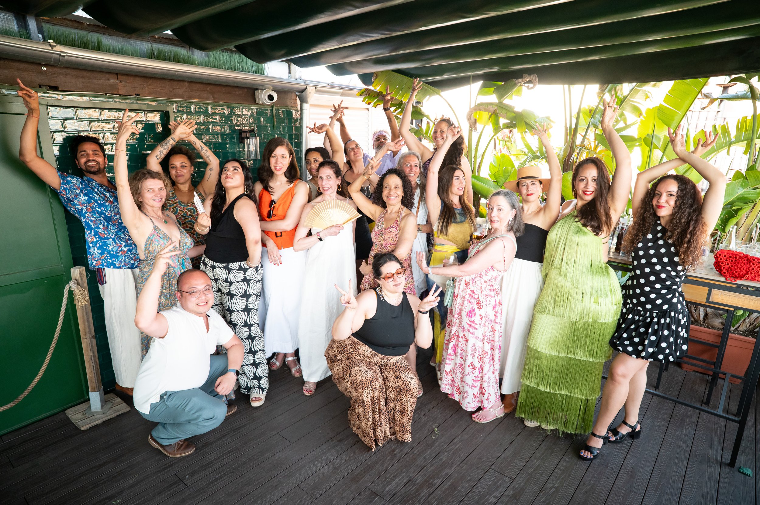 Group of people celebrating at a party or gathering, posing for a photo with smiles and expressive gestures, with green plants in the background.
