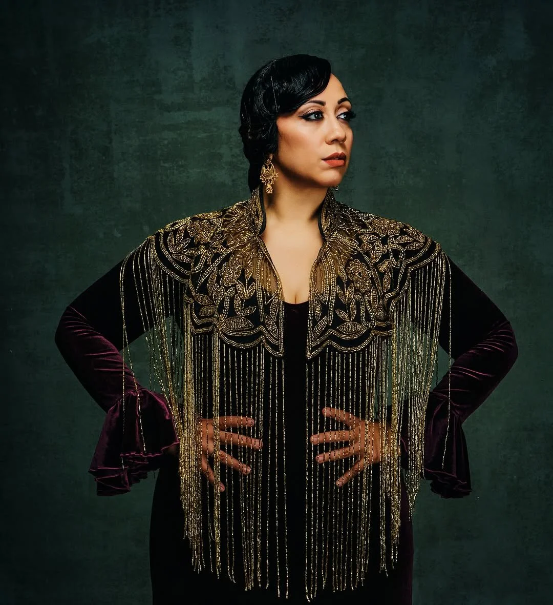 Woman with styled black hair wearing gold embroidered jacket with fringe, black dress, and gold earrings in front of dark green background.