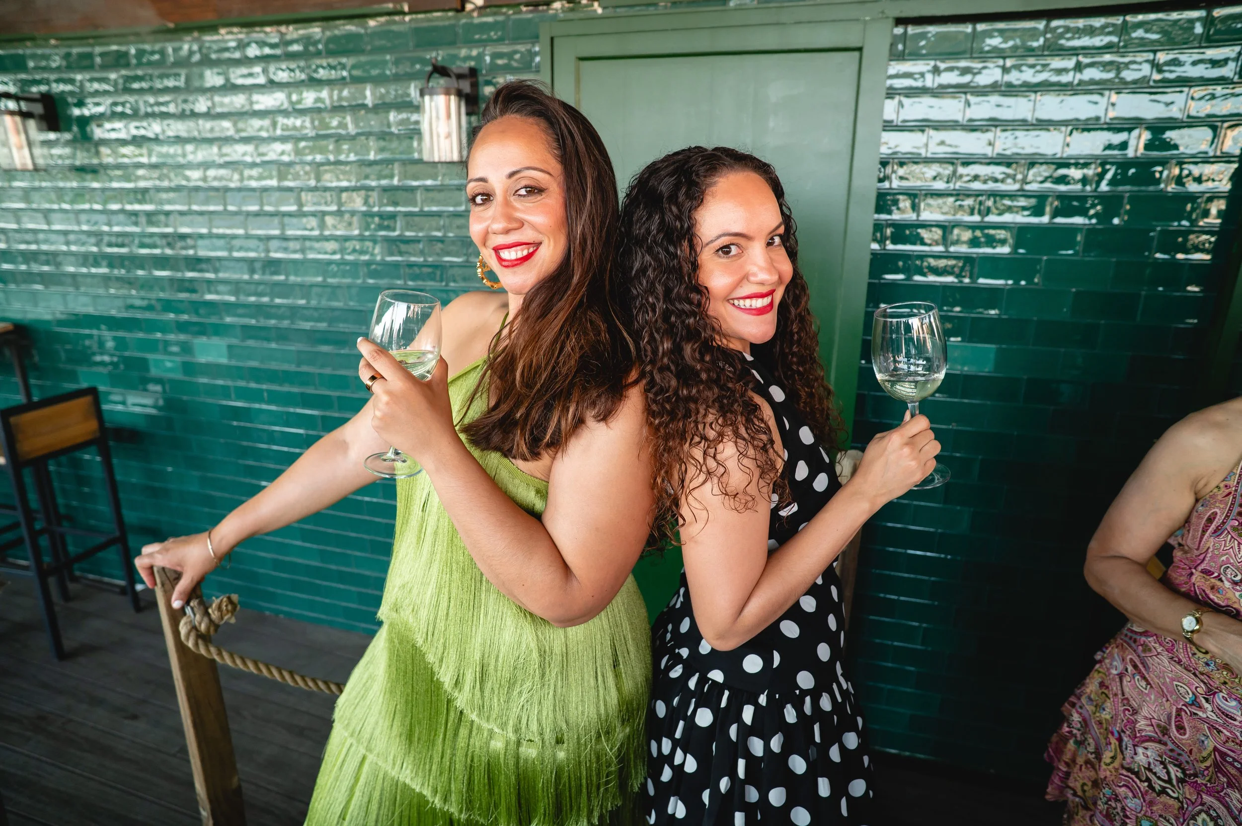 Two women with curly and straight hair in dresses, smiling and holding glasses of white wine at a social gathering against a green tiled wall.