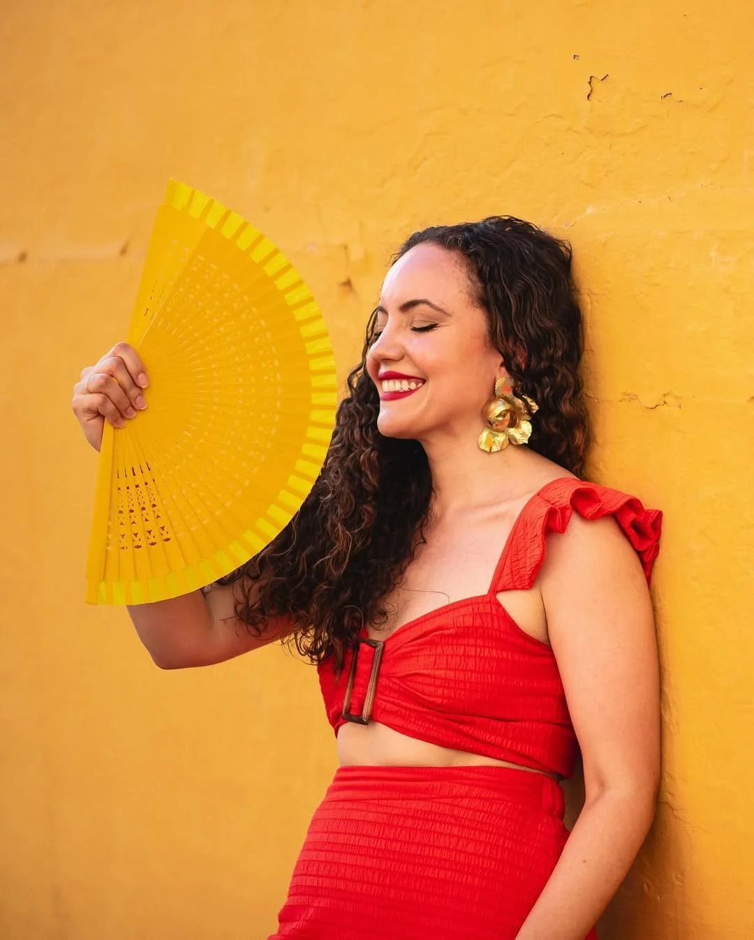 A woman with curly hair and gold earrings wearing a red dress with ruffle shoulder details, smiling with eyes closed, holding a yellow handheld fan, standing against a yellow wall.