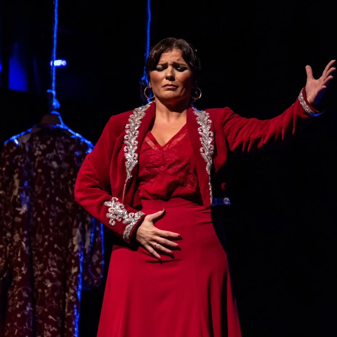 A woman in a red dress and red blazer with silver embroidery is performing on stage, with her eyes closed and her right hand raised, in a theatrical or dramatic pose.