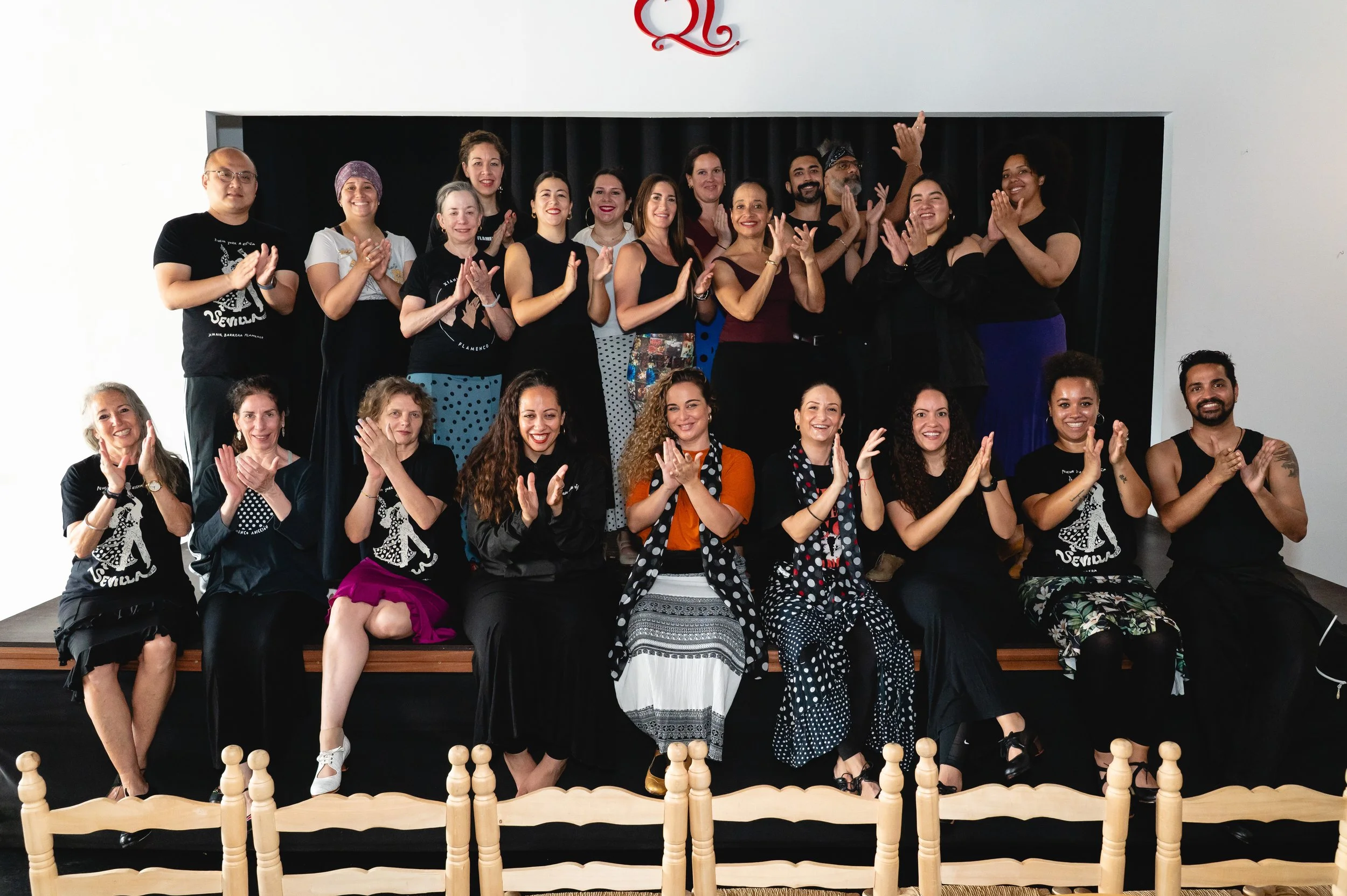 Group of people posing for a photo on a stage, smiling and clapping, with a black curtain as background.