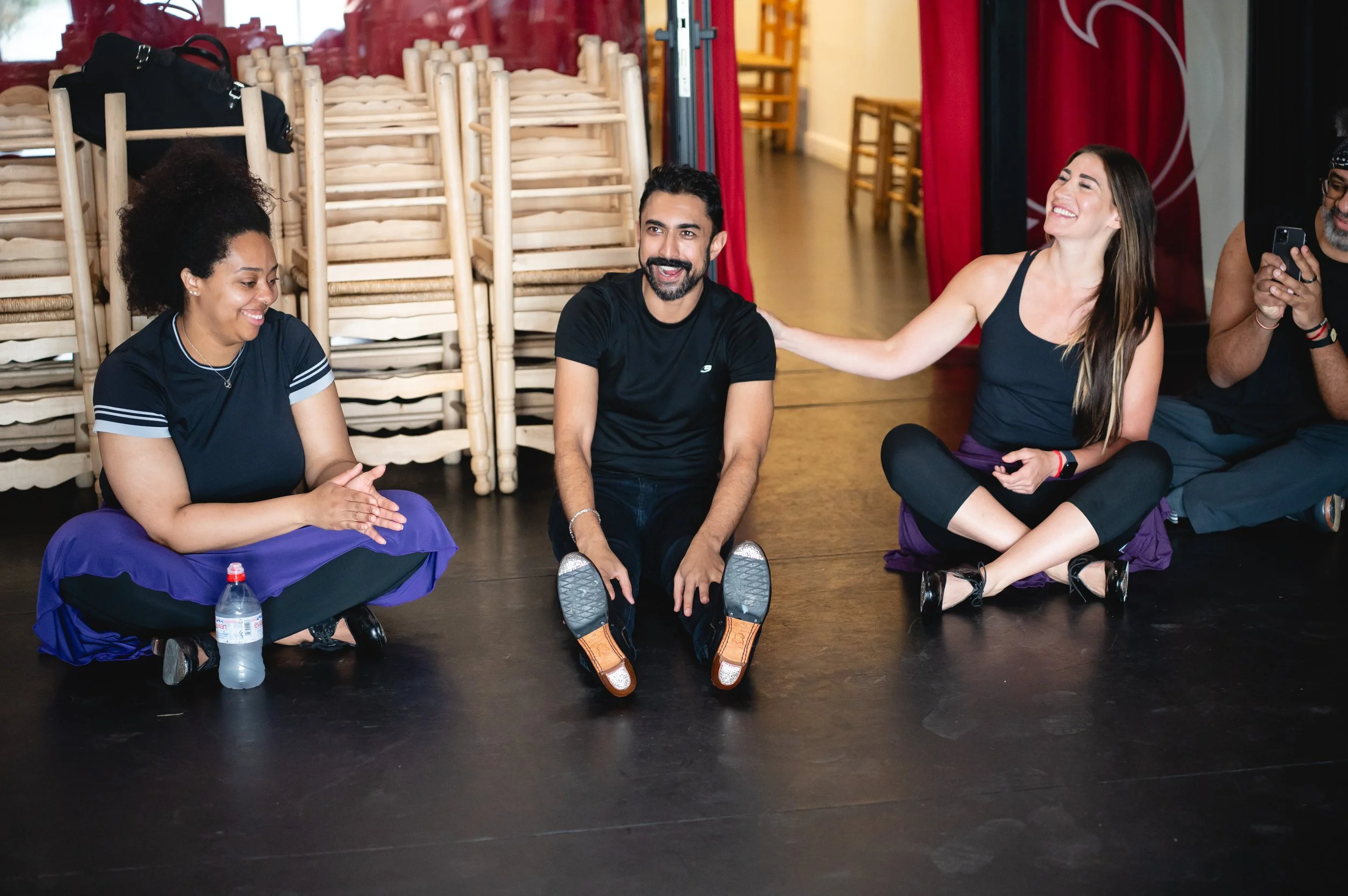 Group of four people sitting cross-legged on the floor, engaging in conversation and smiling, with stacked chairs in the background.