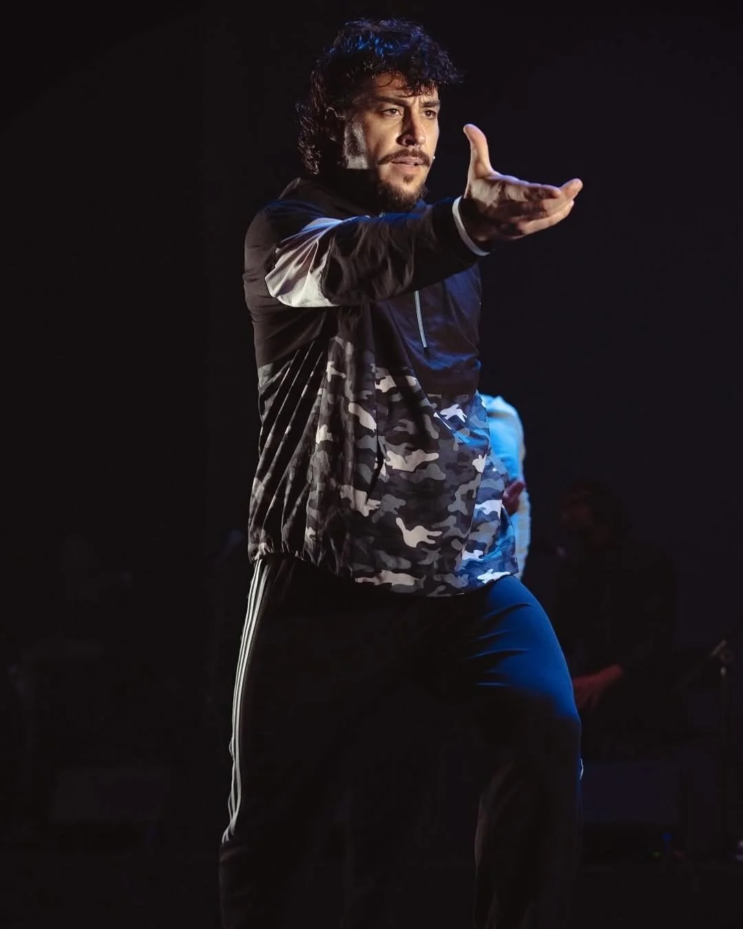 A man with curly hair and a beard, wearing a black and camouflage jacket and black pants, is performing a dance move on stage with his arm extended and hand open.