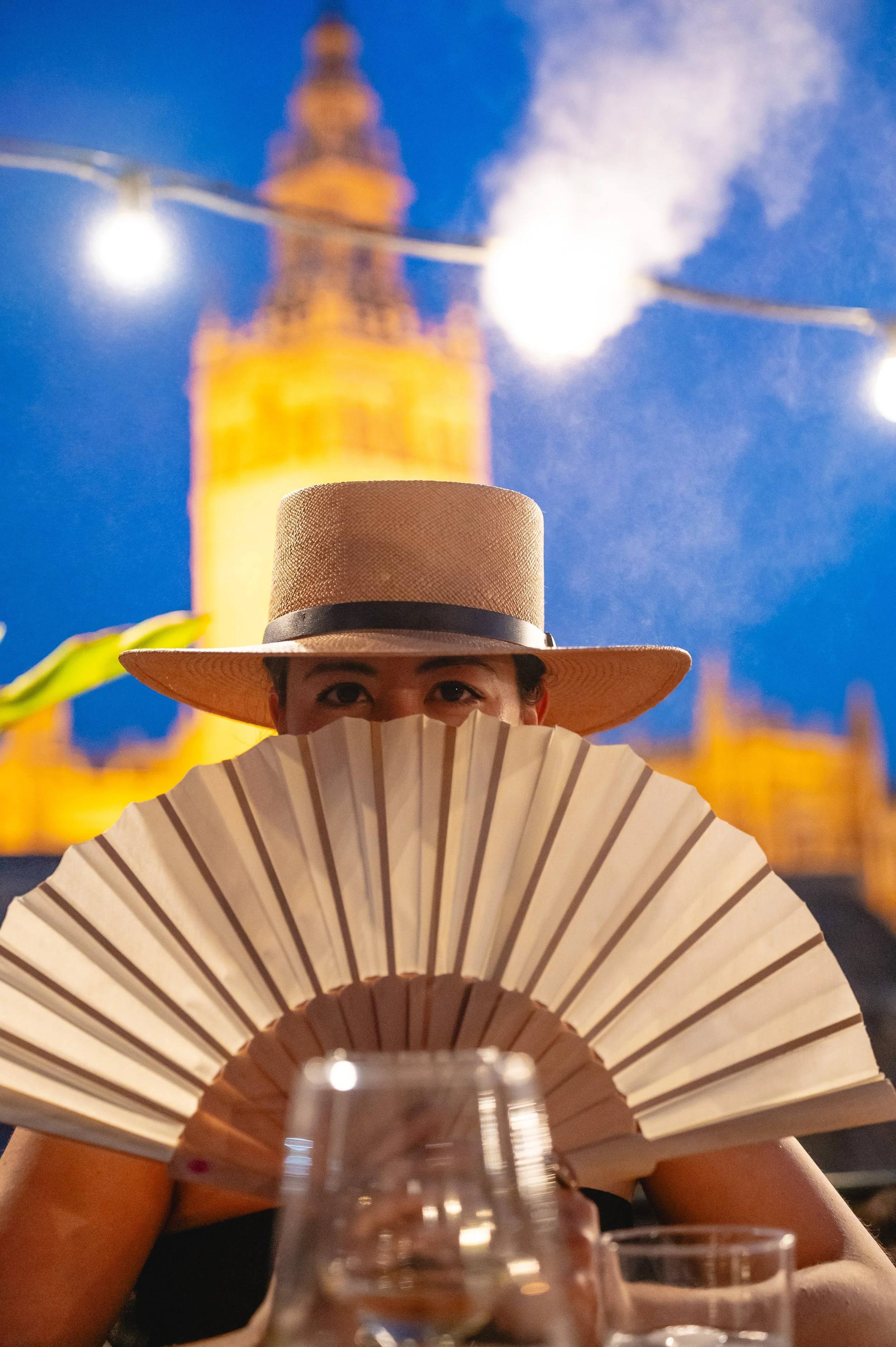 A woman wearing a wide-brimmed straw hat and holding a folding fan, sitting at a table with glasses, in front of a blurred illuminated building and string lights at dusk.