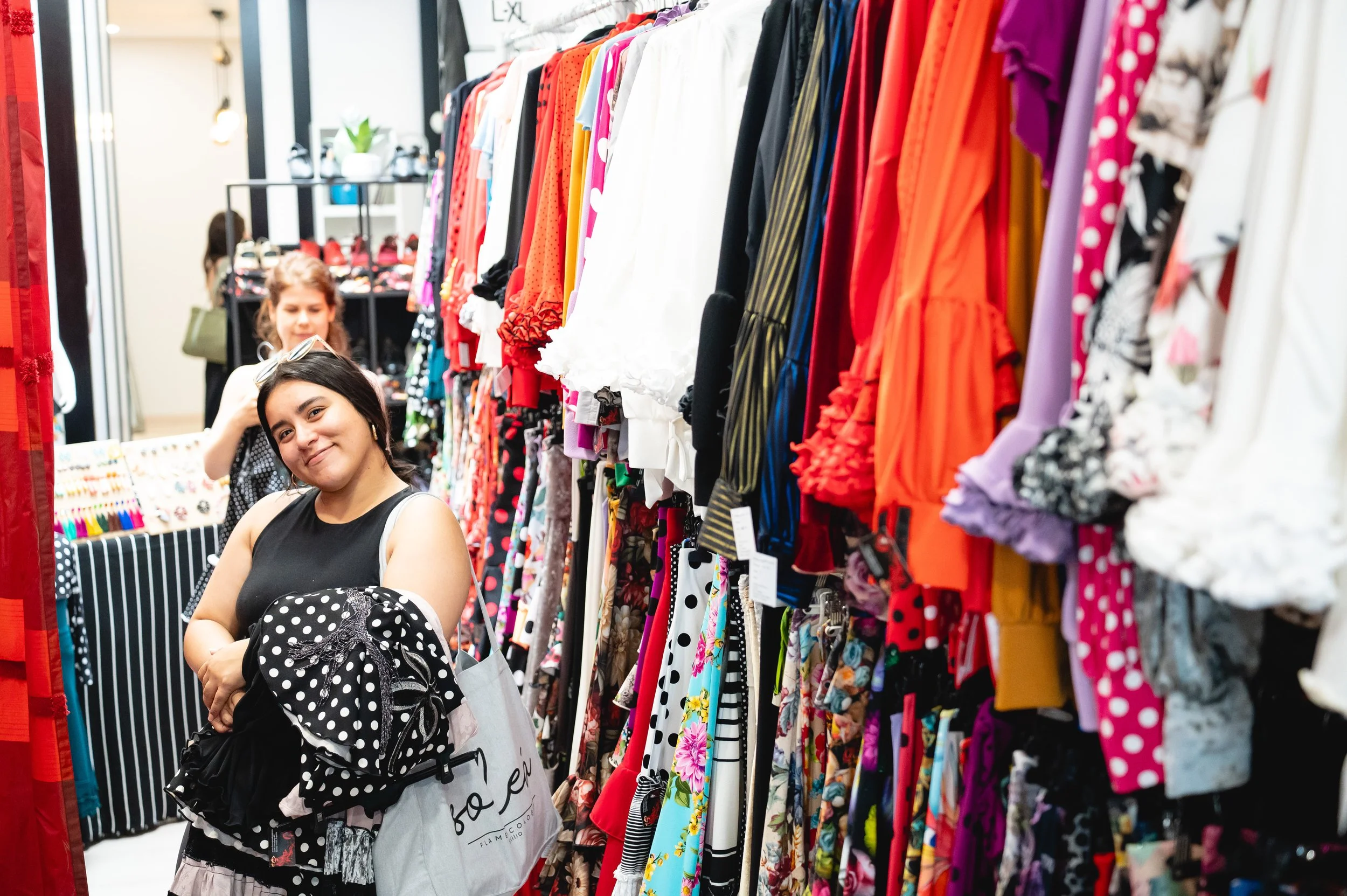 Young woman shopping at a clothing store, holding several polka dot clothing items, with colorful dresses hanging on racks in the background.