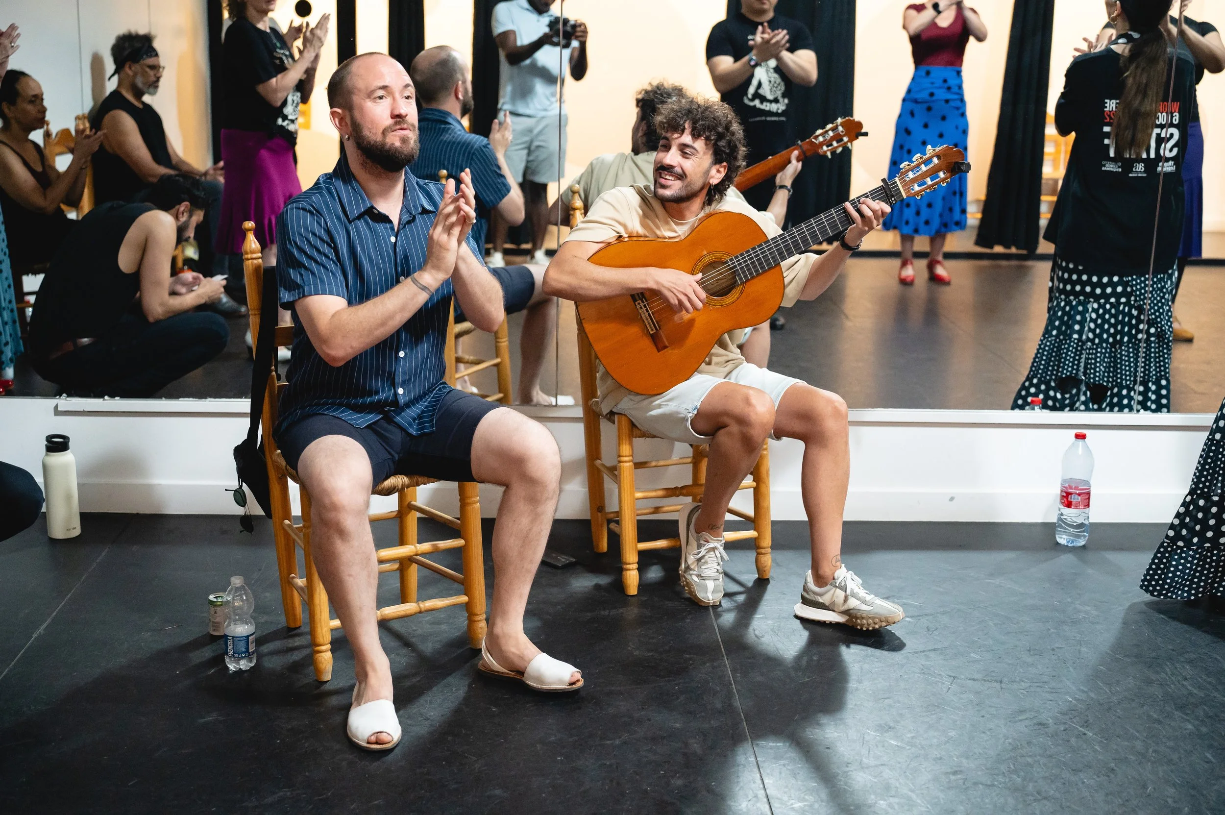 Two men sitting on chairs in front of a mirror, one clapping and the other playing guitar, with a group of people in the background clapping and taking photos.