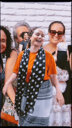 Group of smiling women outdoors against a white brick wall, with one woman in the foreground wearing sunglasses, an orange shirt, and a black and white polka dot scarf.