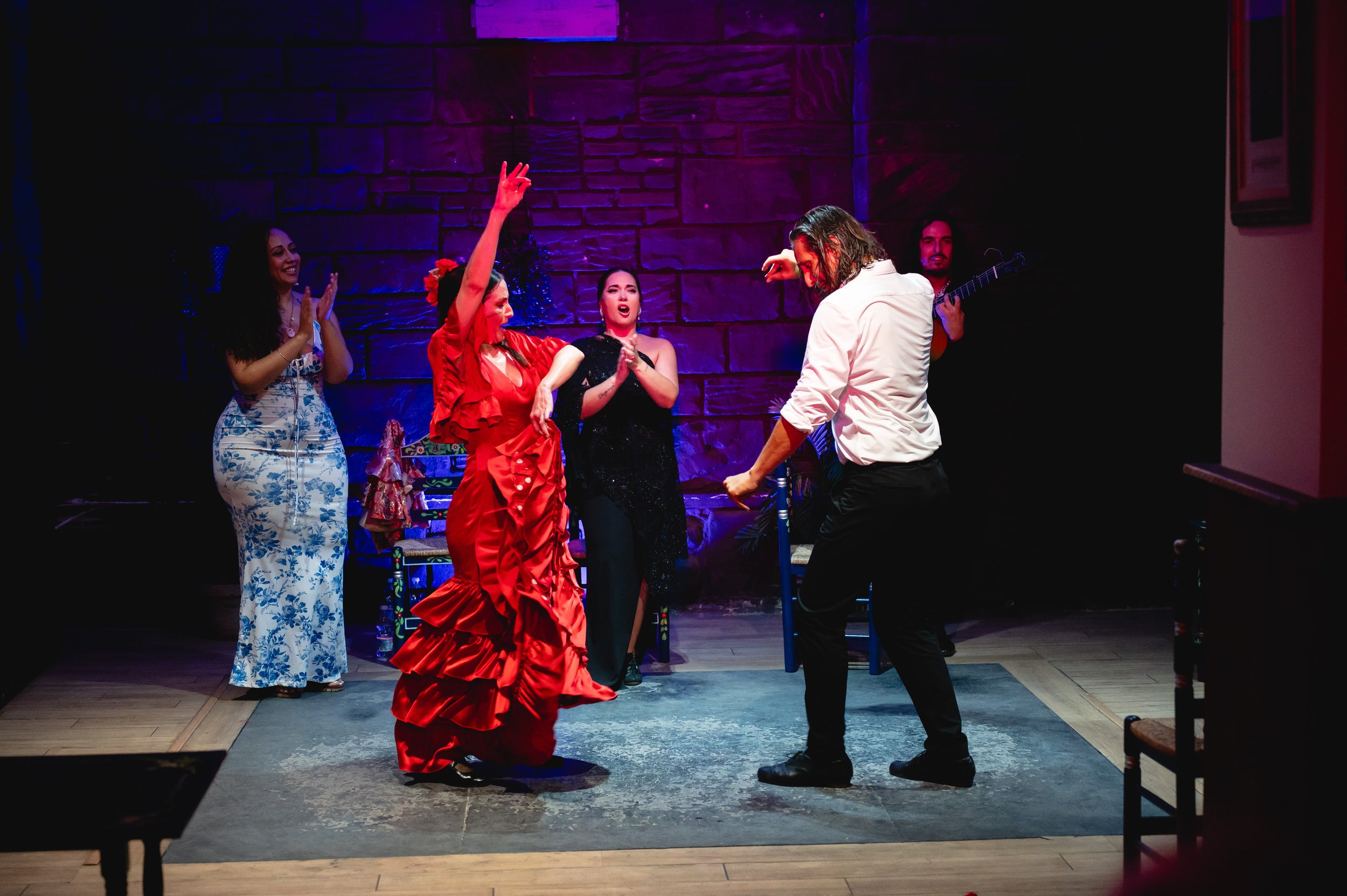 A flamenco dancer in a red dress dances with a man in a white shirt, surrounded by three women clapping and a guitarist playing in the background on a stage with purple lighting.