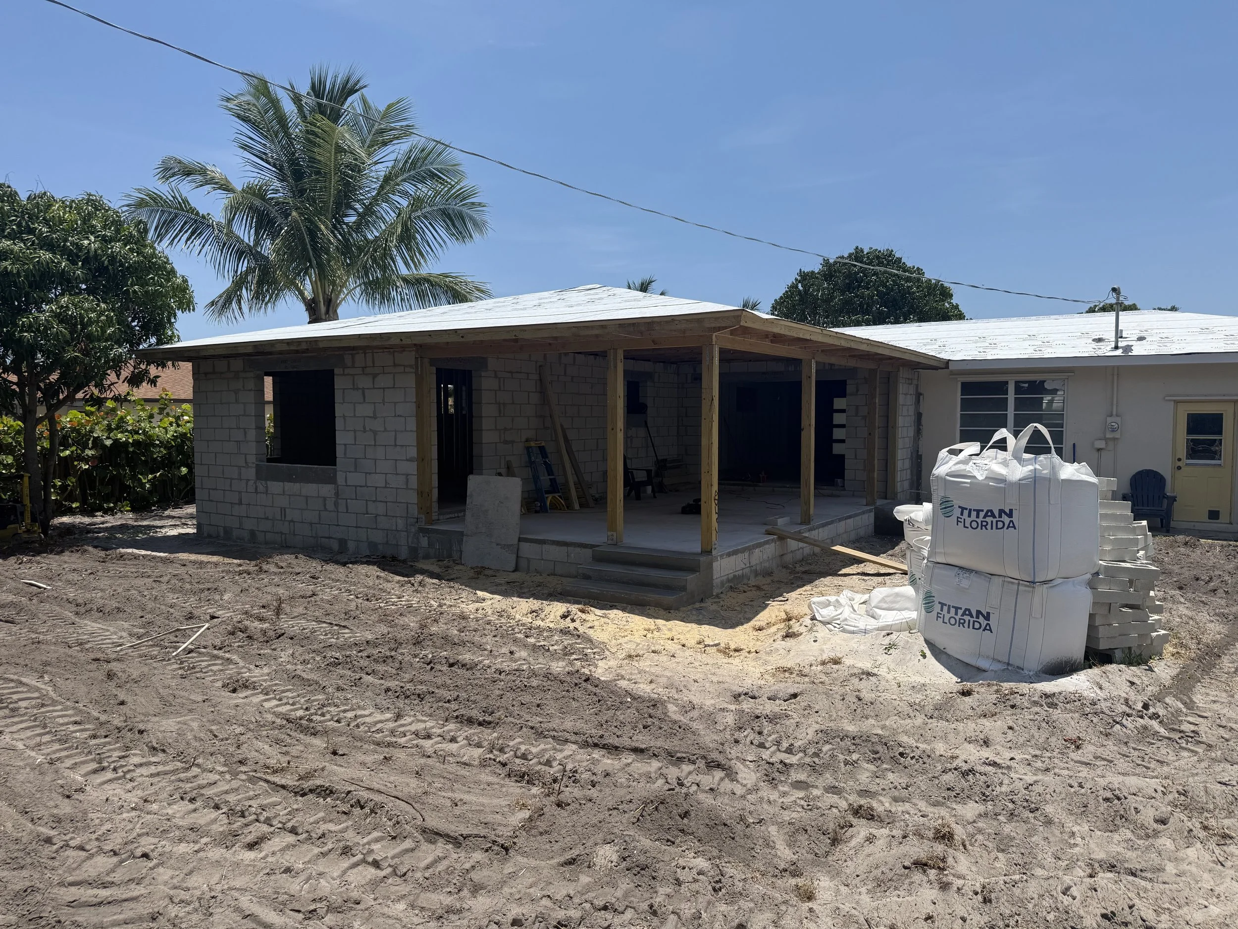 Construction site of a small building with a concrete block exterior and a covered porch, surrounded by dirt and construction materials, under a clear blue sky with palm trees in the background.