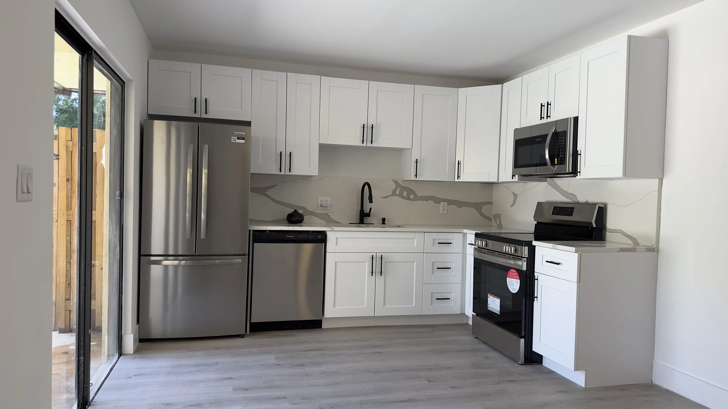 Modern white kitchen with stainless steel appliances, black fixtures, marble backsplash, and light wood flooring.
