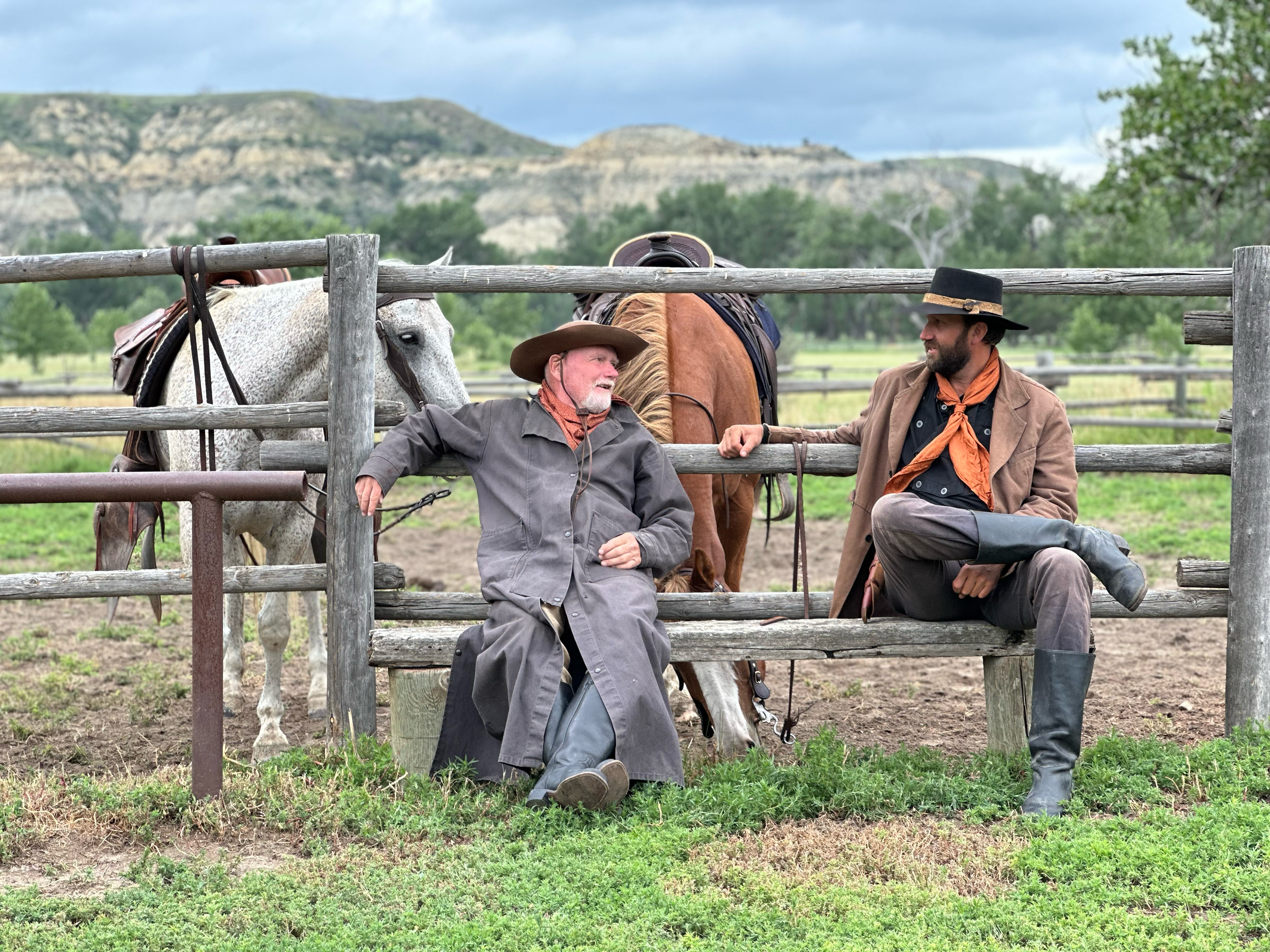 Two cowboys sit by a fence.