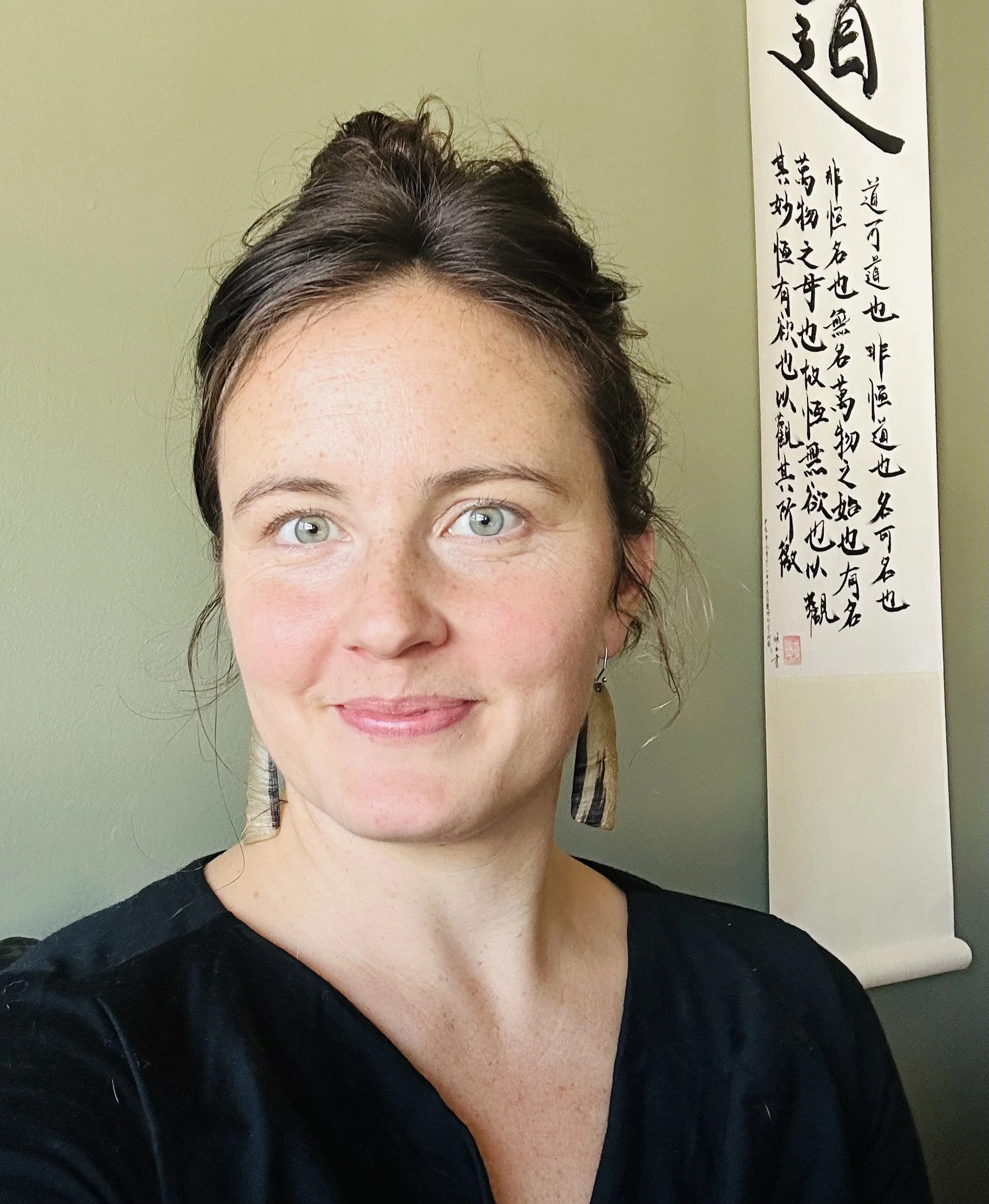 Close-up portrait of a woman with light skin, blue eyes, and dark brown hair tied up, wearing a black top and earrings, standing in front of a green wall with a hanging scroll of Chinese calligraphy.