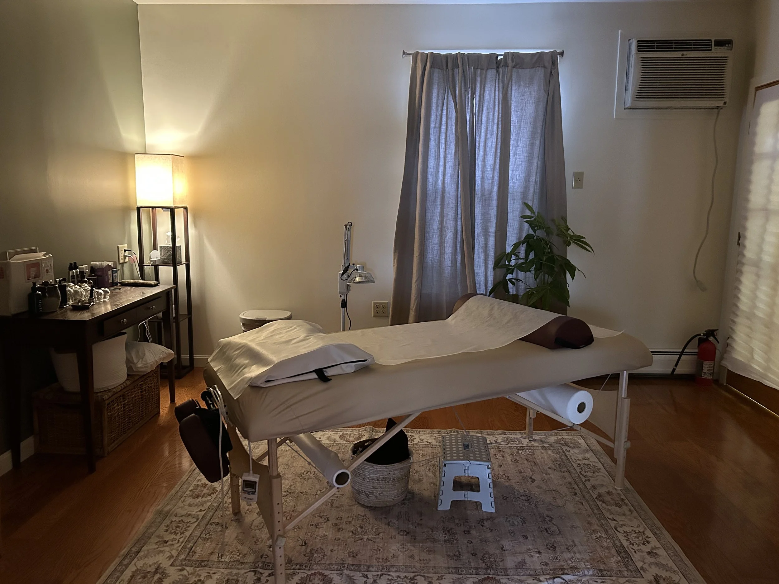 A massage therapy room with a massage table covered with a white sheet, a bolster, a lunging pillow, and a face cradle. There is a wooden side table with bottles, cups, and a machine. A tall lamp provides warm lighting. A window with curtains, a pott