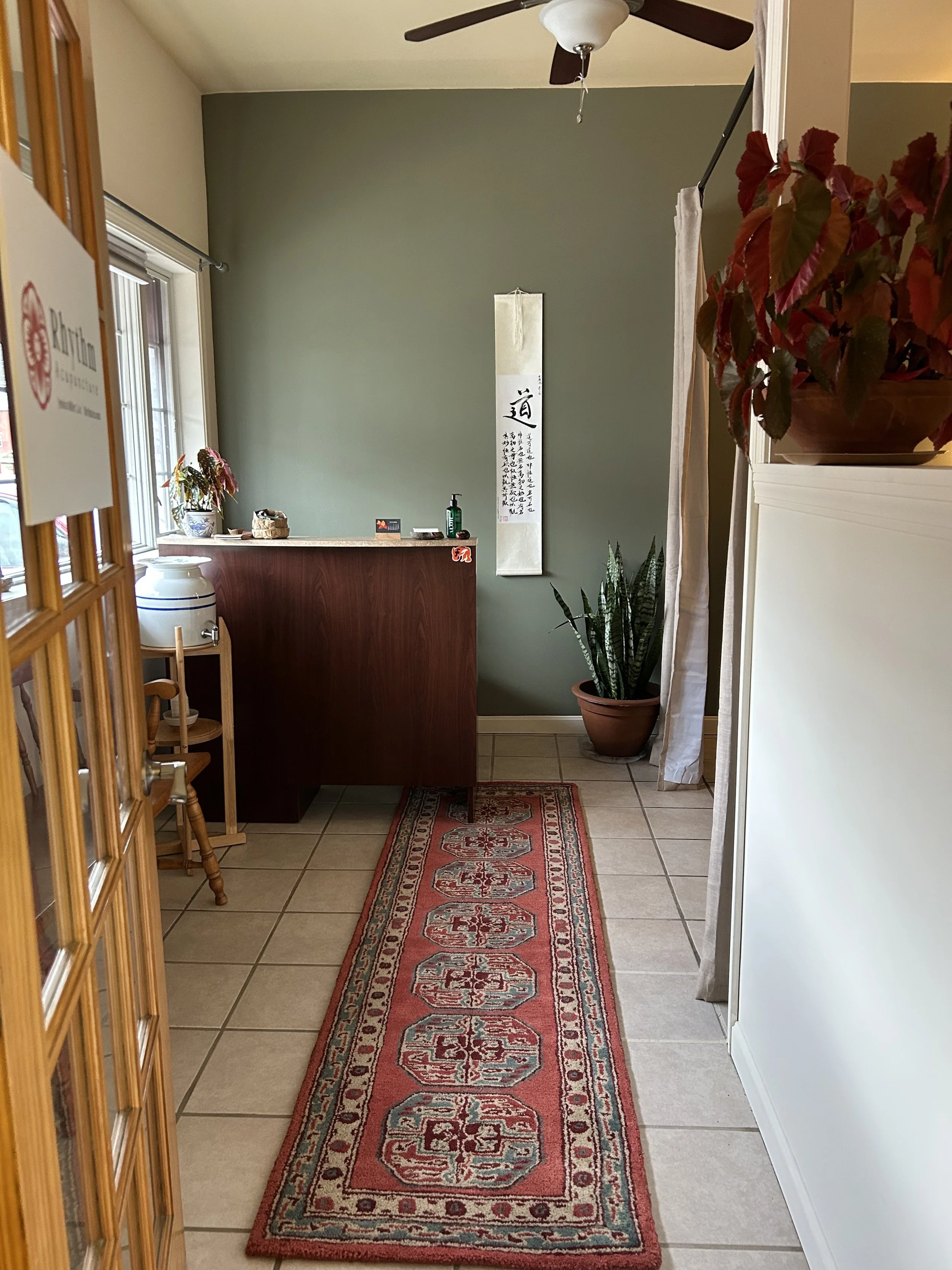 A view of a reception or waiting area with a small red patterned rug on tiled floor, a green accent wall, a potted plant, a hanging scroll with Chinese calligraphy, a hallway curtain, and a ceiling fan.