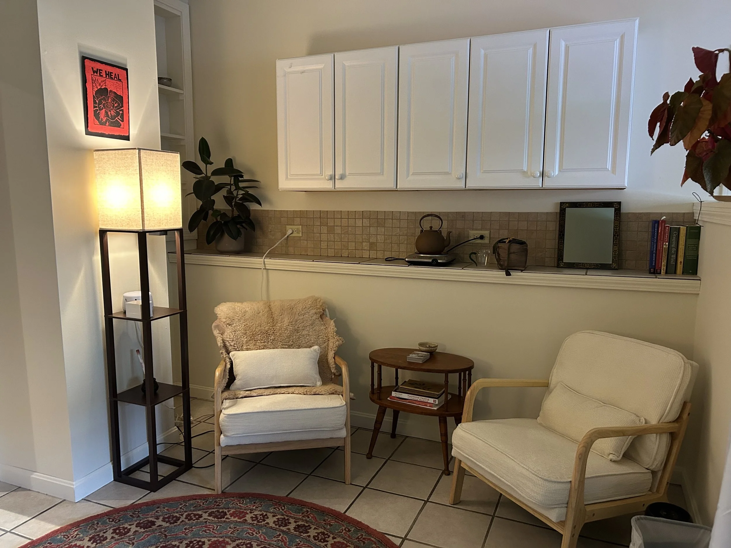 Cozy living corner with two armchairs, a small wooden side table, a tall floor lamp, and decorative items, set against a kitchen area with white cabinets and a tiled backsplash.