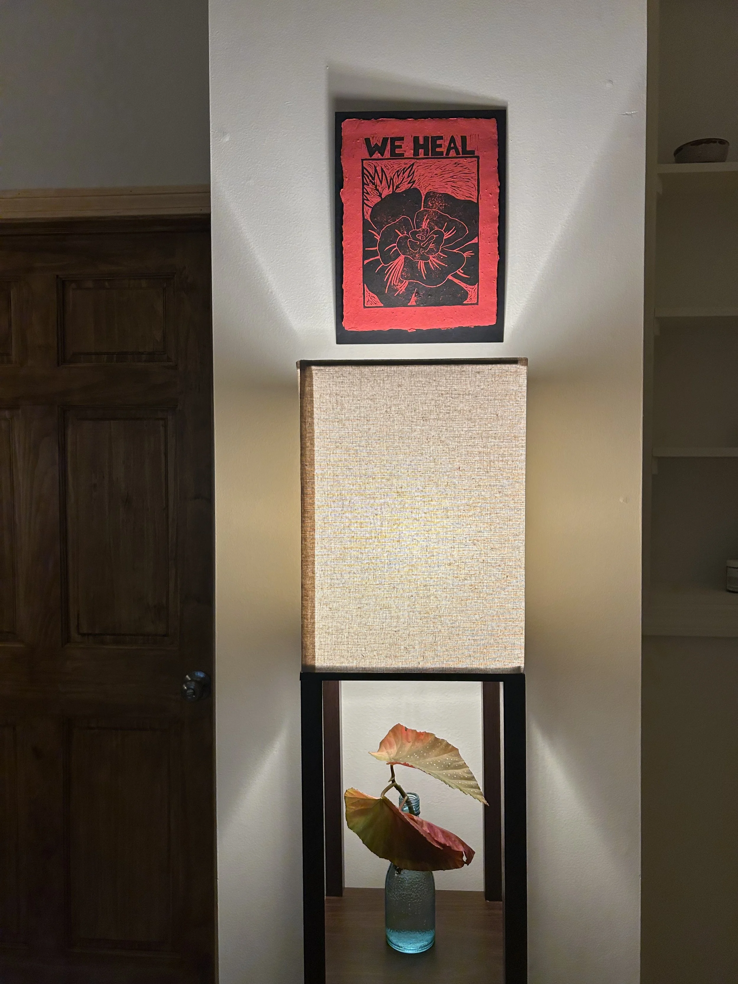 Decorative room corner with a piece of artwork, a lamp with a fabric shade, and a vase with large leaves.