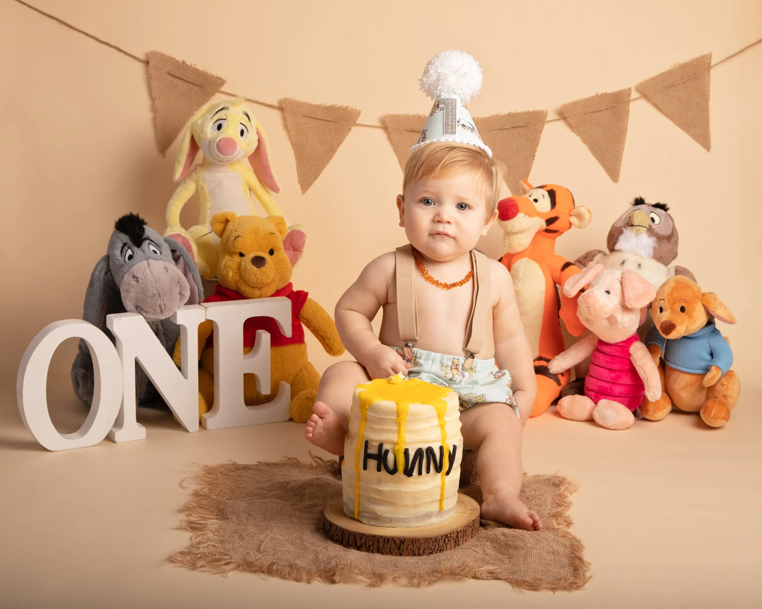 A 1 year old boy celebrating his birthday with a cake, wearing a birthday hat, surrounded by stuffed animals including Winnie the Pooh, Tigger, and Eeyore. The boy is holding a cake with a "HAPPY BIRTHDAY" message and has a serious expression.
