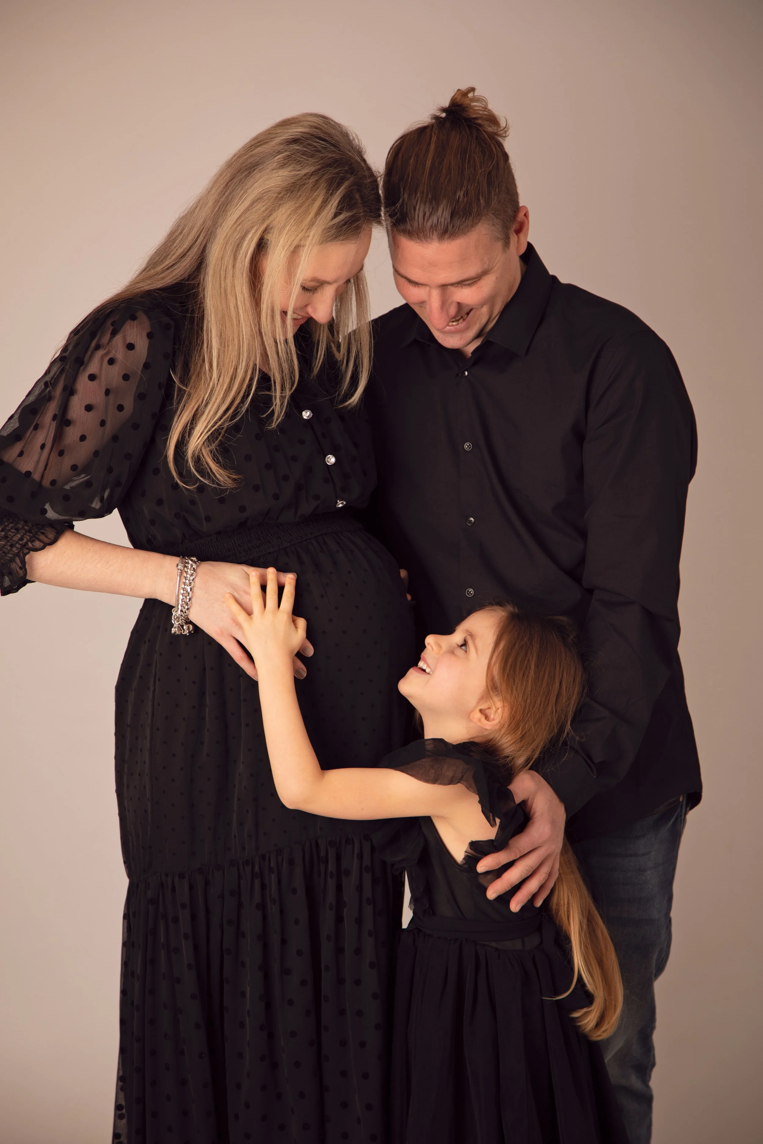 A happy group of three people, including a pregnant woman, smiling and touching her baby bump while looking at a young girl, all dressed in black.