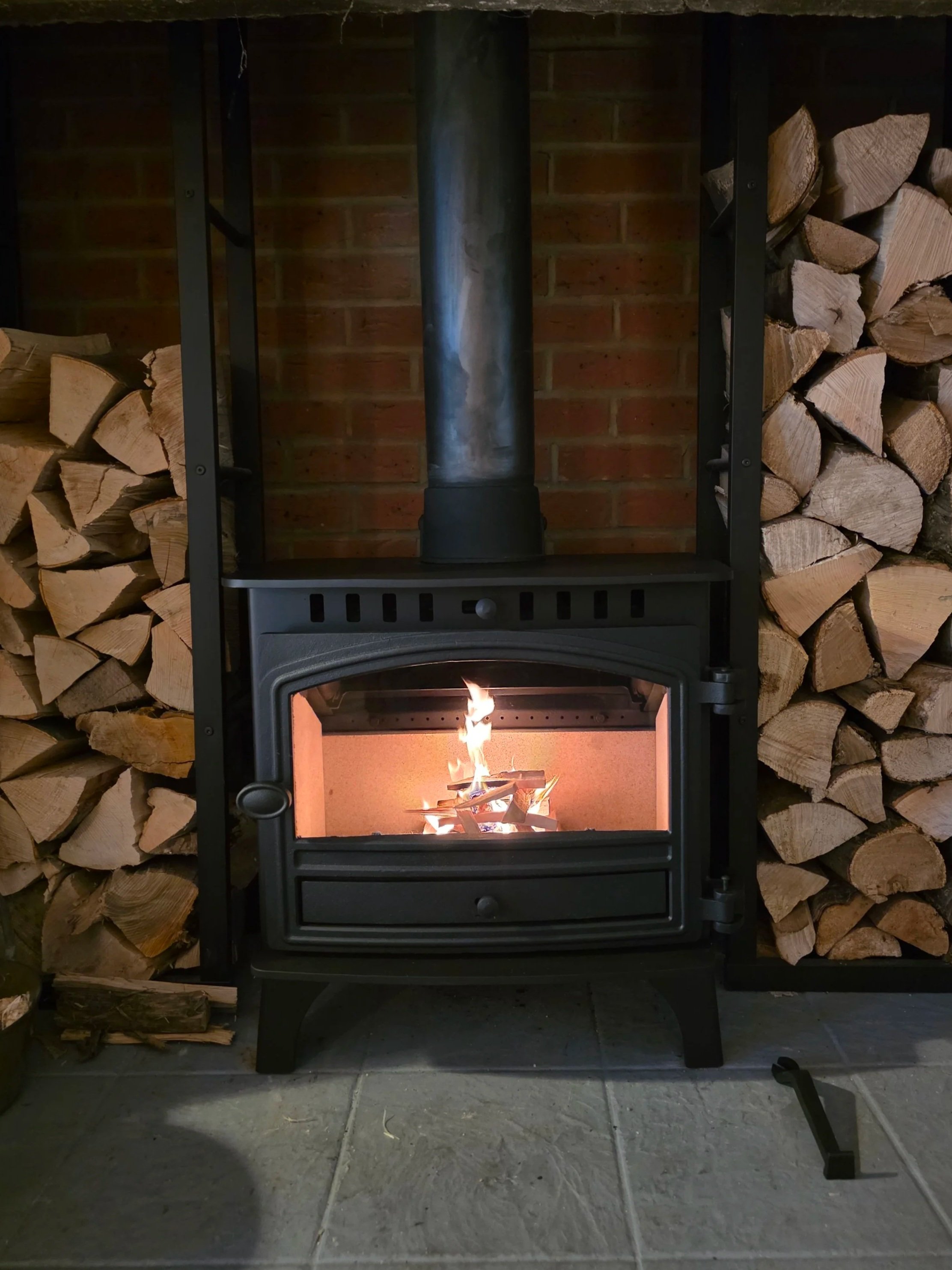 A wood-burning stove with a fire inside, placed on a tiled floor against a brick wall, with stacked firewood on either side.