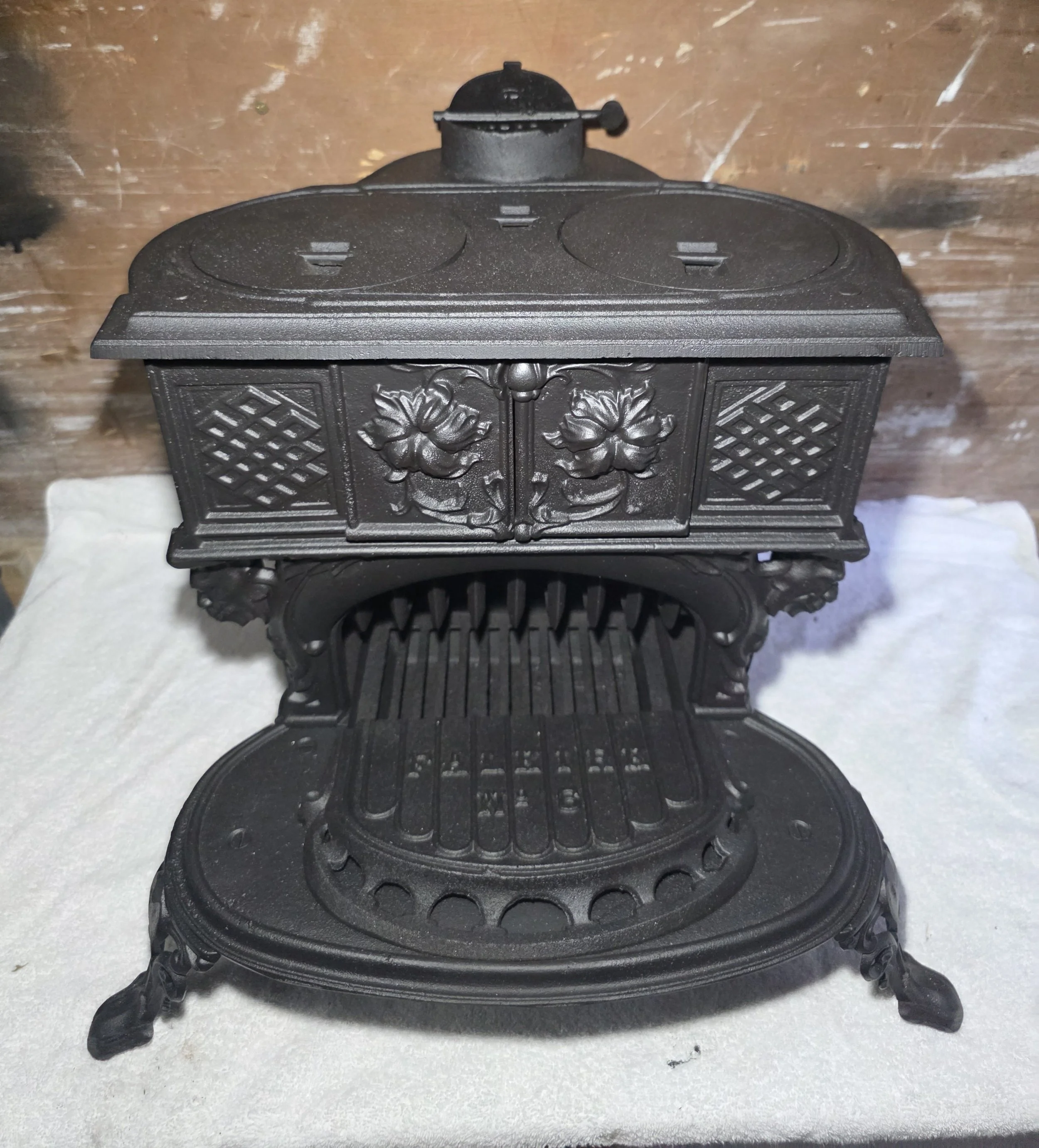 A vintage cast iron stove from 1920 fully refurbished with decorative floral and lattice designs on the doors, featuring three circular burners on top and ornate legs, set on a snowy surface with a wooden background.