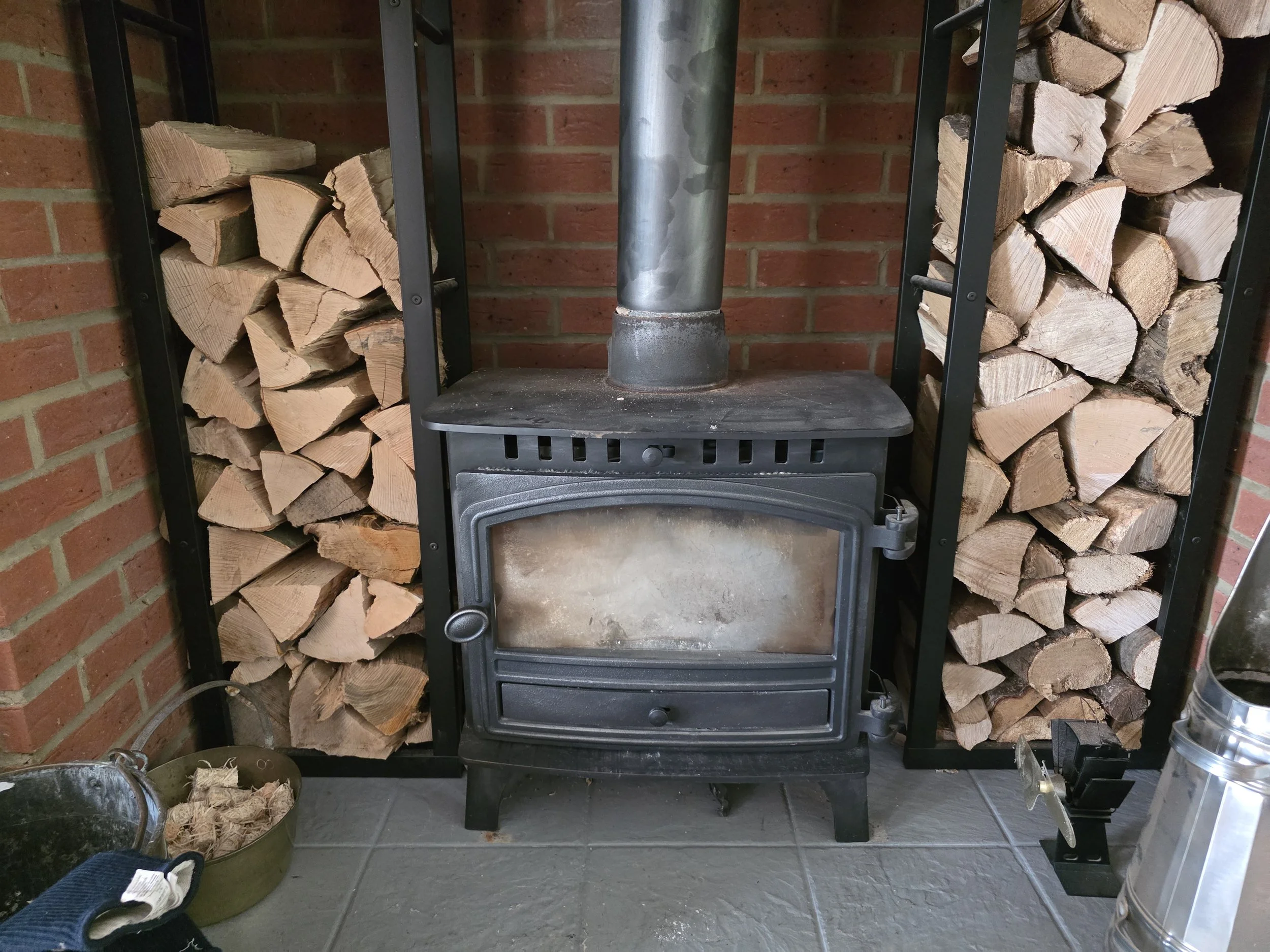 Stacked firewood on either side of a black wood stove with a metal pipe extending from top to the brick wall behind. Metal tools and a pot are visible on the floor. herald hunter 8