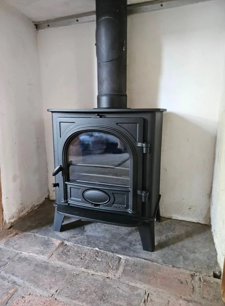 A black wood stove with a glass door, metal handle, and chimney pipe in a corner of a room with brick and plaster walls.